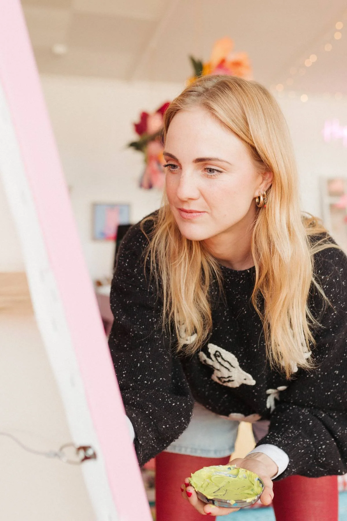 Young woman in black sweater with white design, holding decorated cupcake, leaning forward in a room with pink and floral decorations.