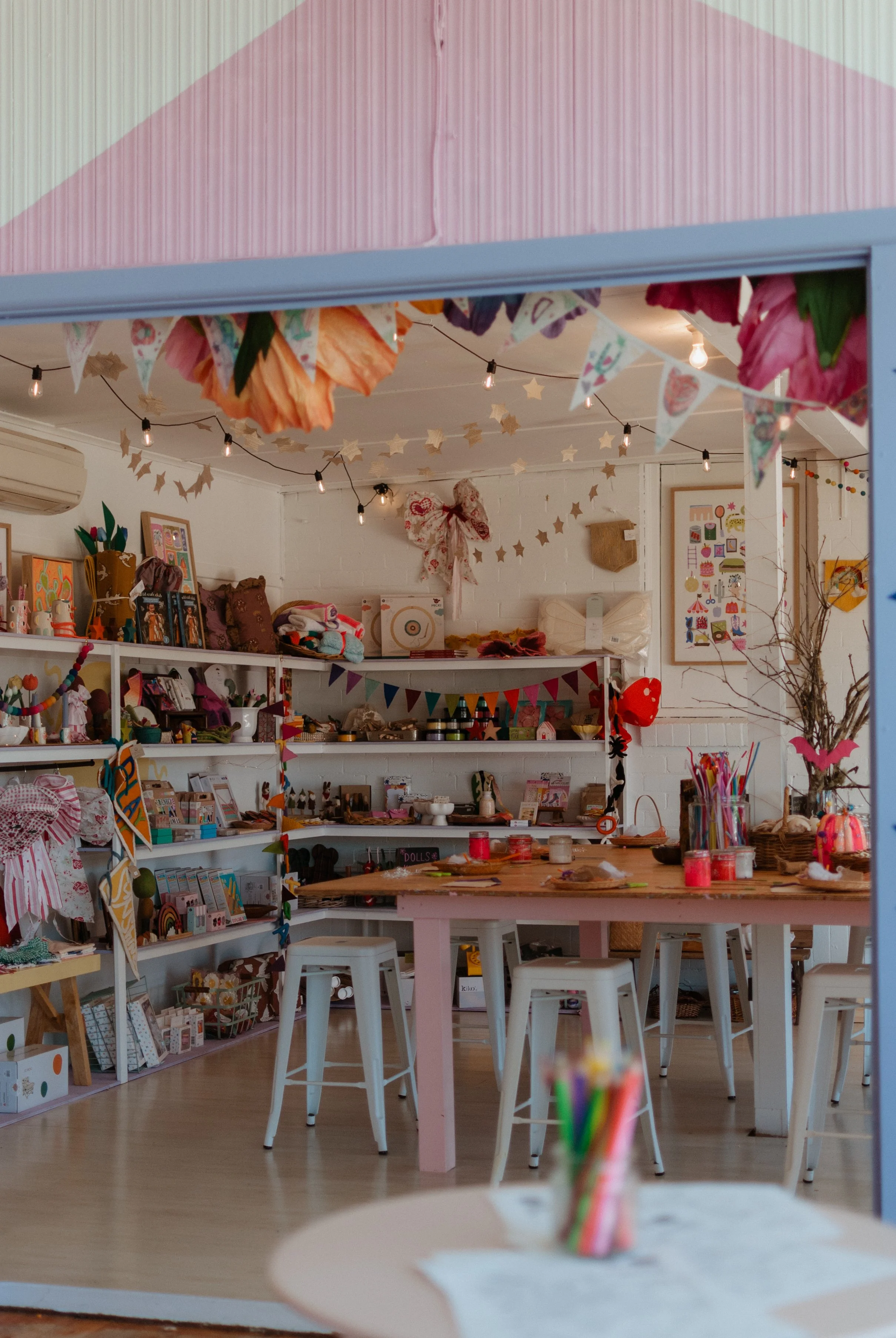 Decorated craft or gift shop with shelves of art supplies, wall art, hanging paper flowers, and festive bunting; table with colored markers and paper in foreground.