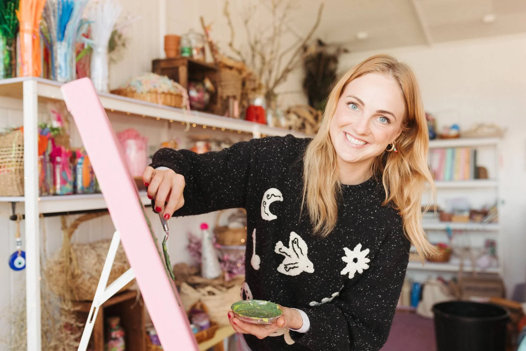 A woman with blonde hair and blue eyes smiling while painting on an easel in a craft room, holding a small bowl of green paint.