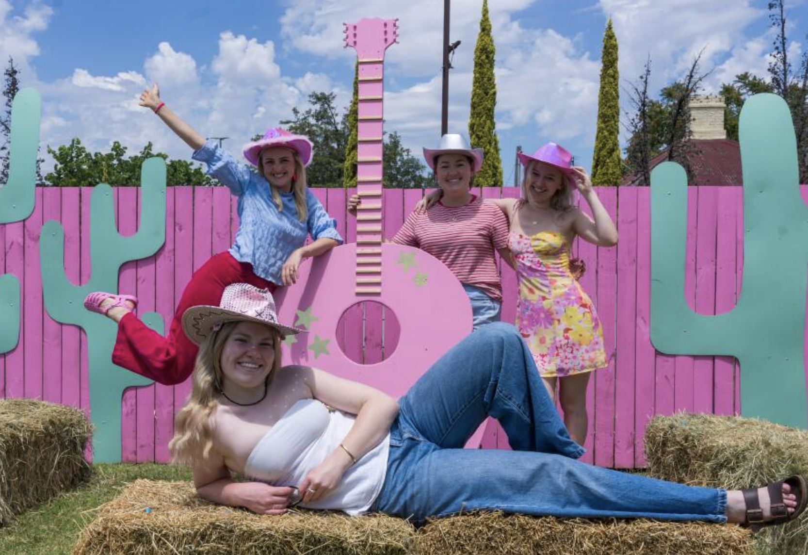 Group of five girls dressed in summer clothes and pink cowboy hats, posing in front of a pink and teal cactus-themed backdrop at an outdoor event.