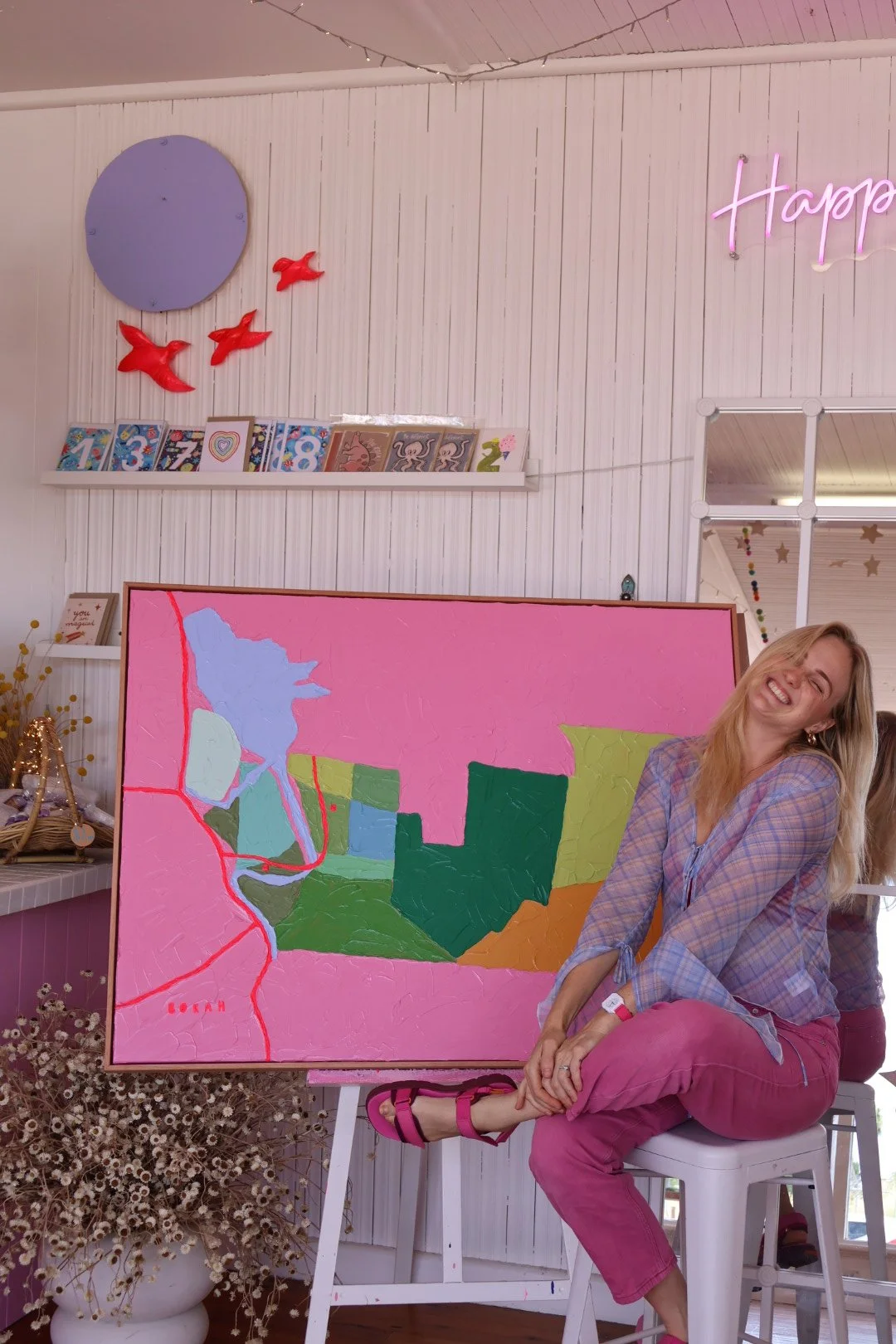 A smiling woman with long blonde hair sitting on a white stool in front of a pink and green abstract painting in a gallery or art space. The background has white paneled walls, a string of fairy lights, and a neon sign that says 'Happy.' There are shelves with children's books and decorative items, and a plant with small white flowers in the foreground.