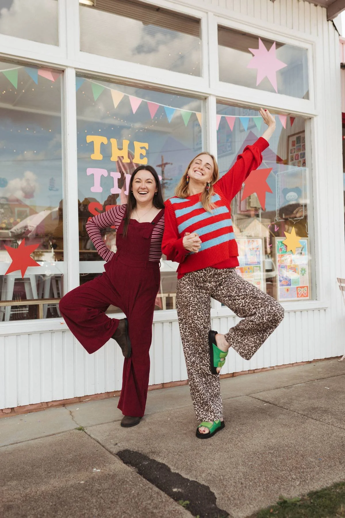 Two women standing outside a colorful storefront, smiling and raising their arms. One woman is dressed in red overalls and a striped shirt, and the other is wearing a red sweater with blue stripes and leopard print pants.