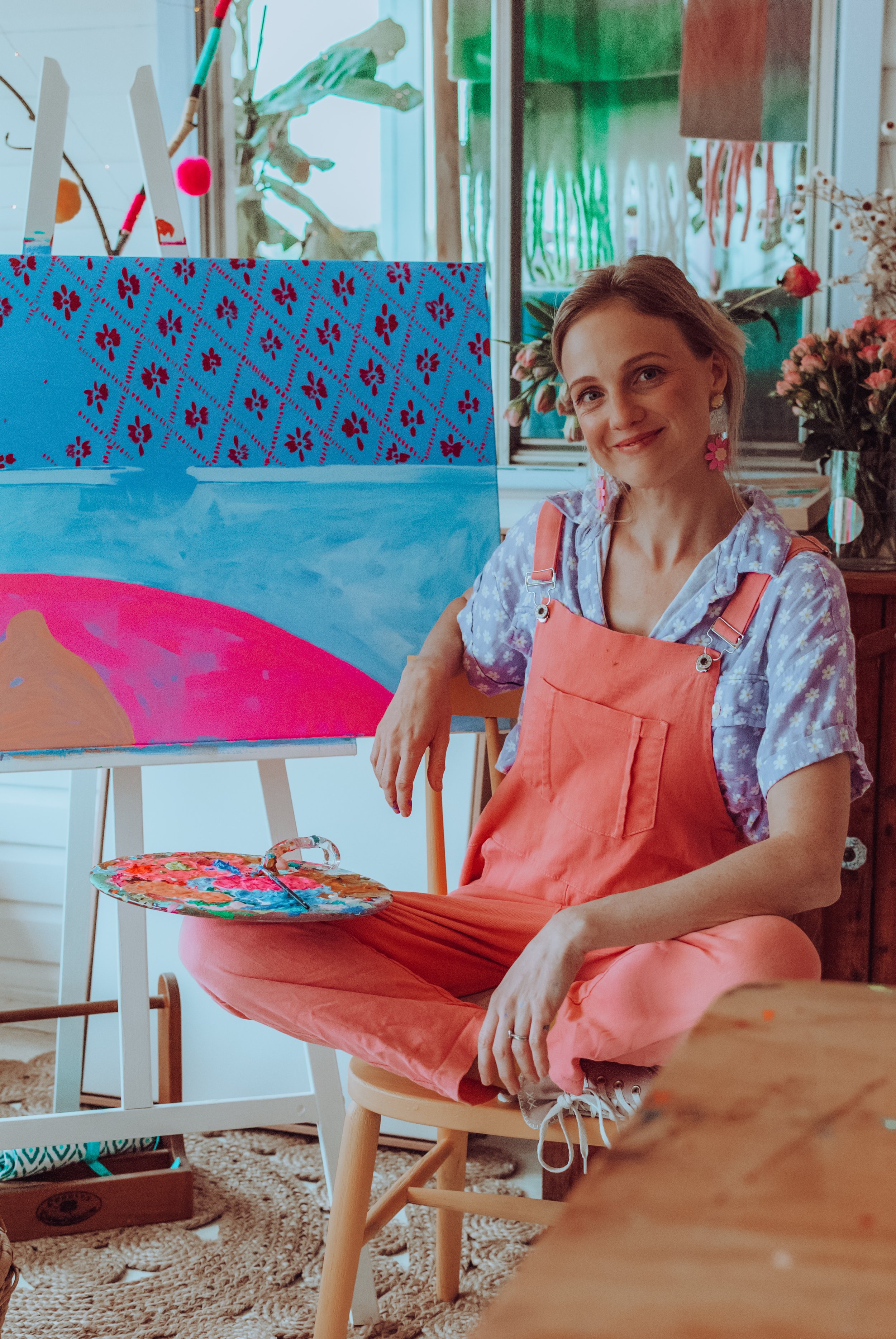 A woman sitting on a wooden stool, wearing pink overalls and a blue floral shirt, holding a painter's palette, with an easel and a colorful painting of a sunset behind her in an art studio surrounded by plants and flowers.