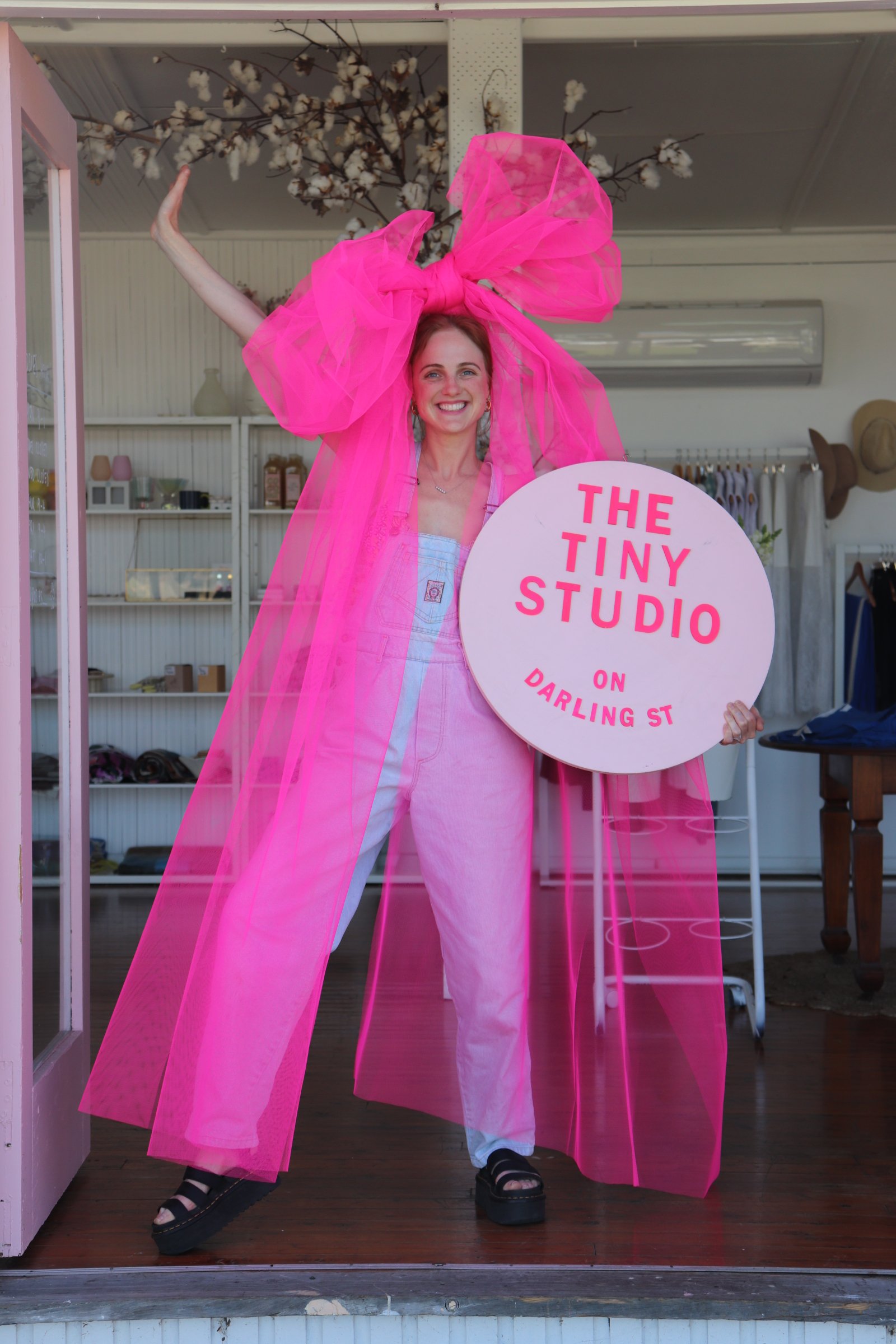 A woman smiling and holding a sign that says 'The Tiny Studio on Darling St'. She is wearing a pink, sheer, oversized bow and matching pink dress, standing inside a boutique or studio with clothing and accessories in the background.