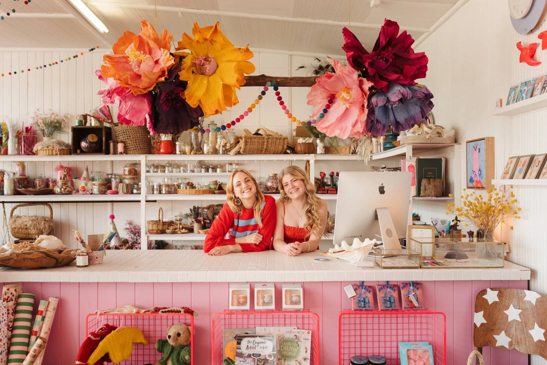 Two young women sitting behind a decorated retail counter in a craft store, smiling at the camera. The counter has a pink front and is topped with craft supplies and a computer. Bright, colorful paper flowers hang from the ceiling above them, and shelves behind them display jars, baskets, and various craft items. The store has a cheerful, creative atmosphere.