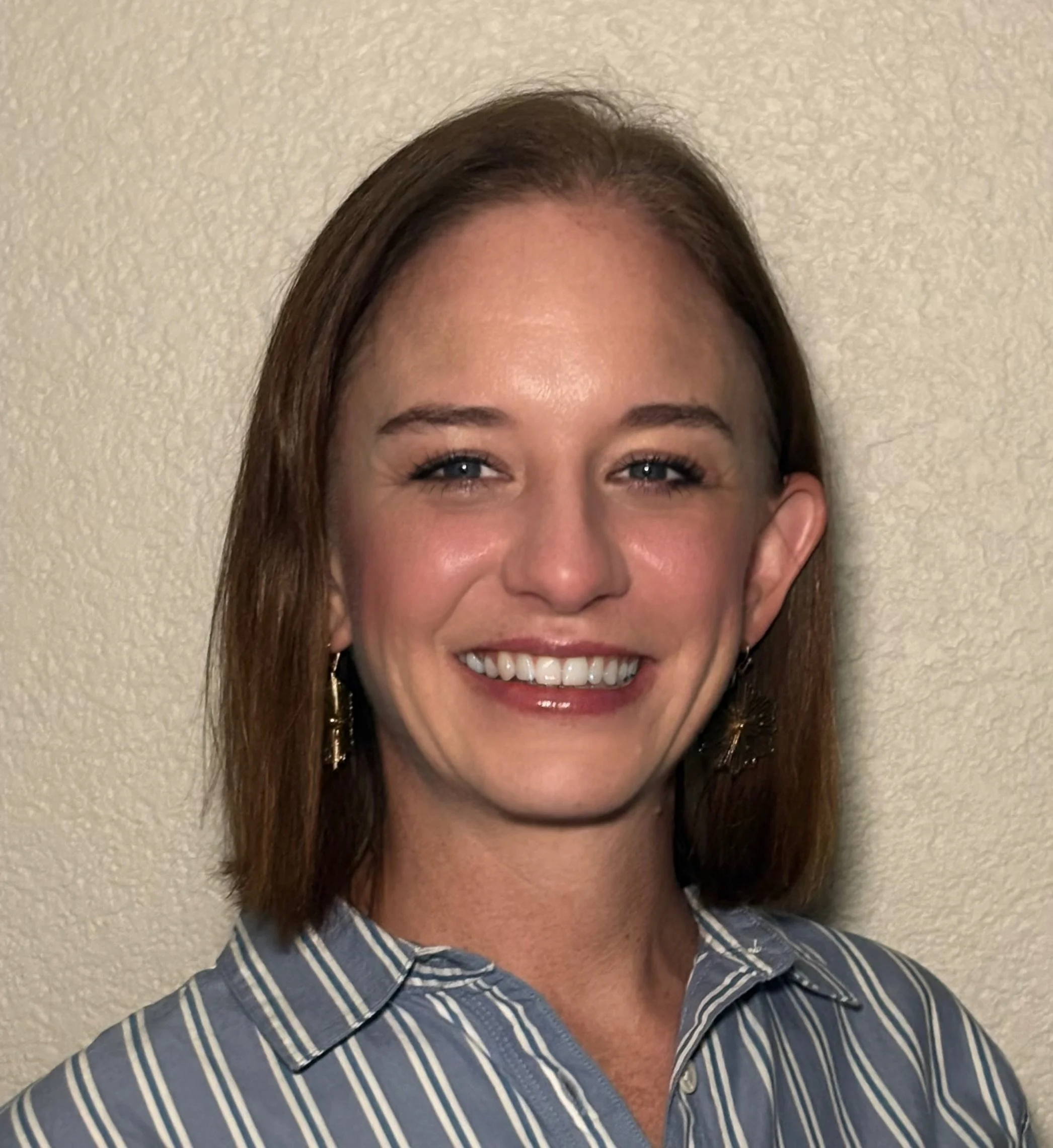 Close-up of a smiling woman, Rachel Hackworth, with brown hair and blue eyes, wearing a striped blue and white shirt, standing against a plain light-colored wall.