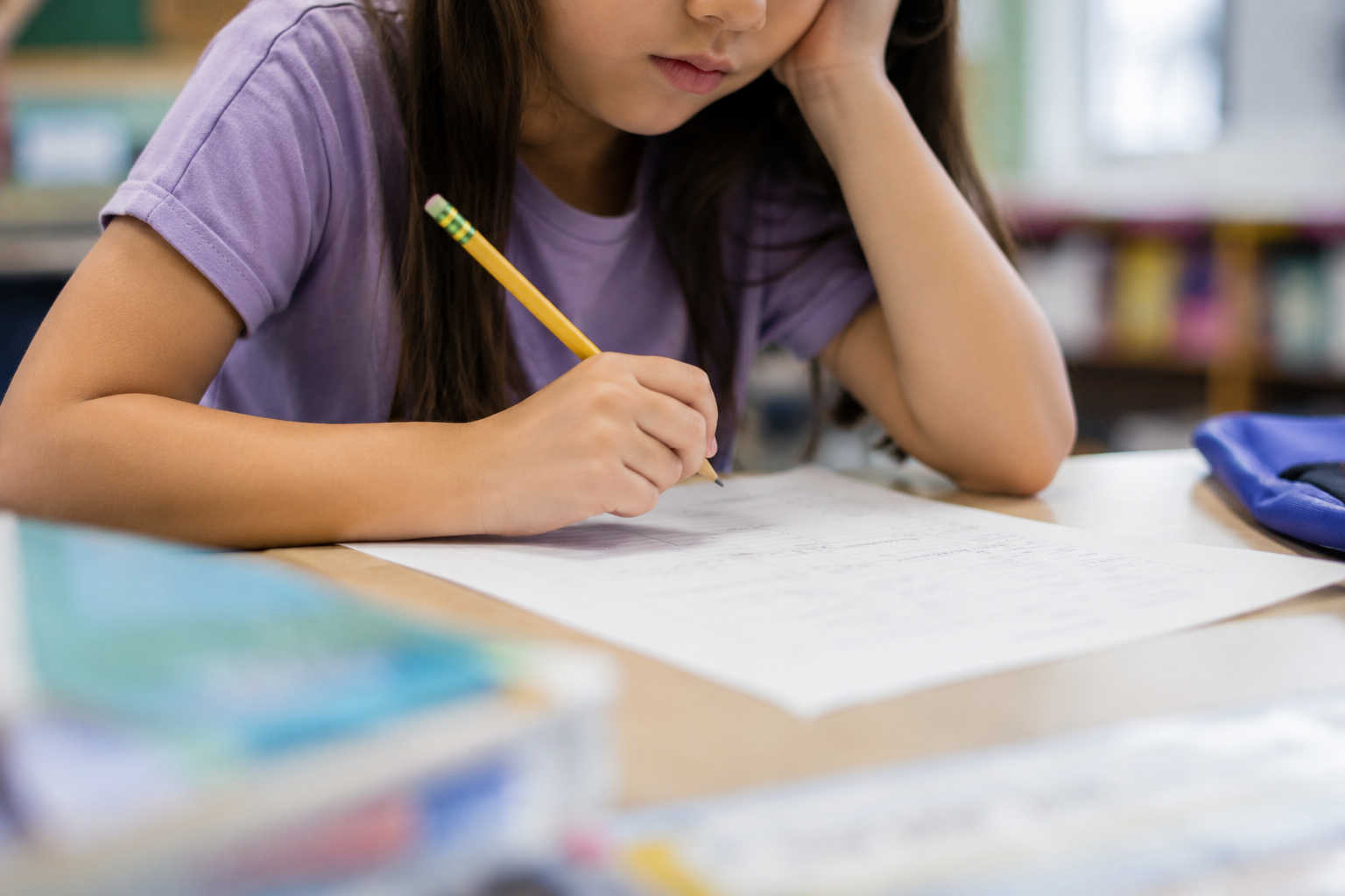 A girl in a purple shirt writing on a paper with a pencil in a classroom.