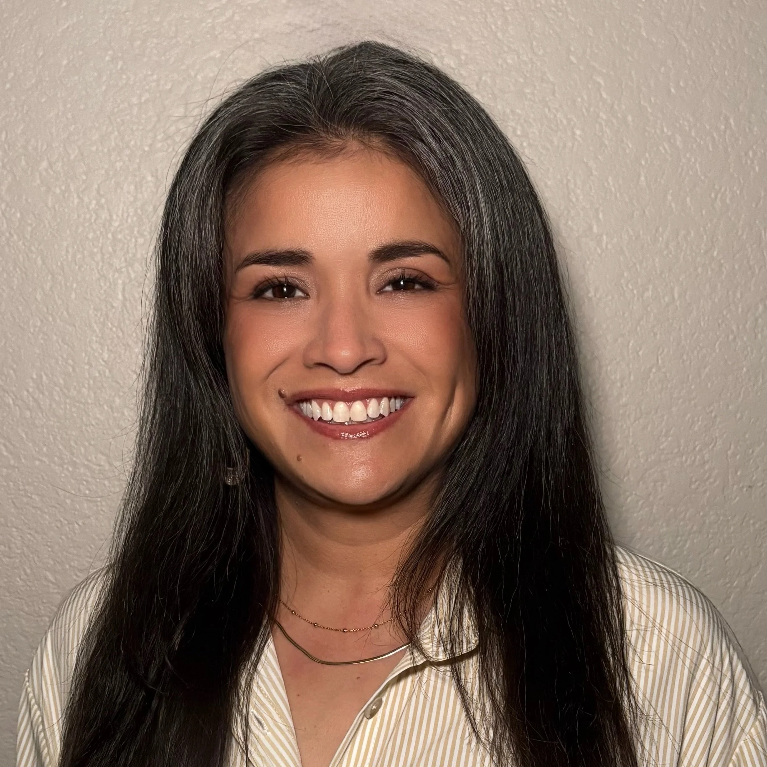 Close-up of a smiling woman, Becky Choat, with long black hair, wearing a gold necklace and a striped shirt, standing against a beige textured wall.