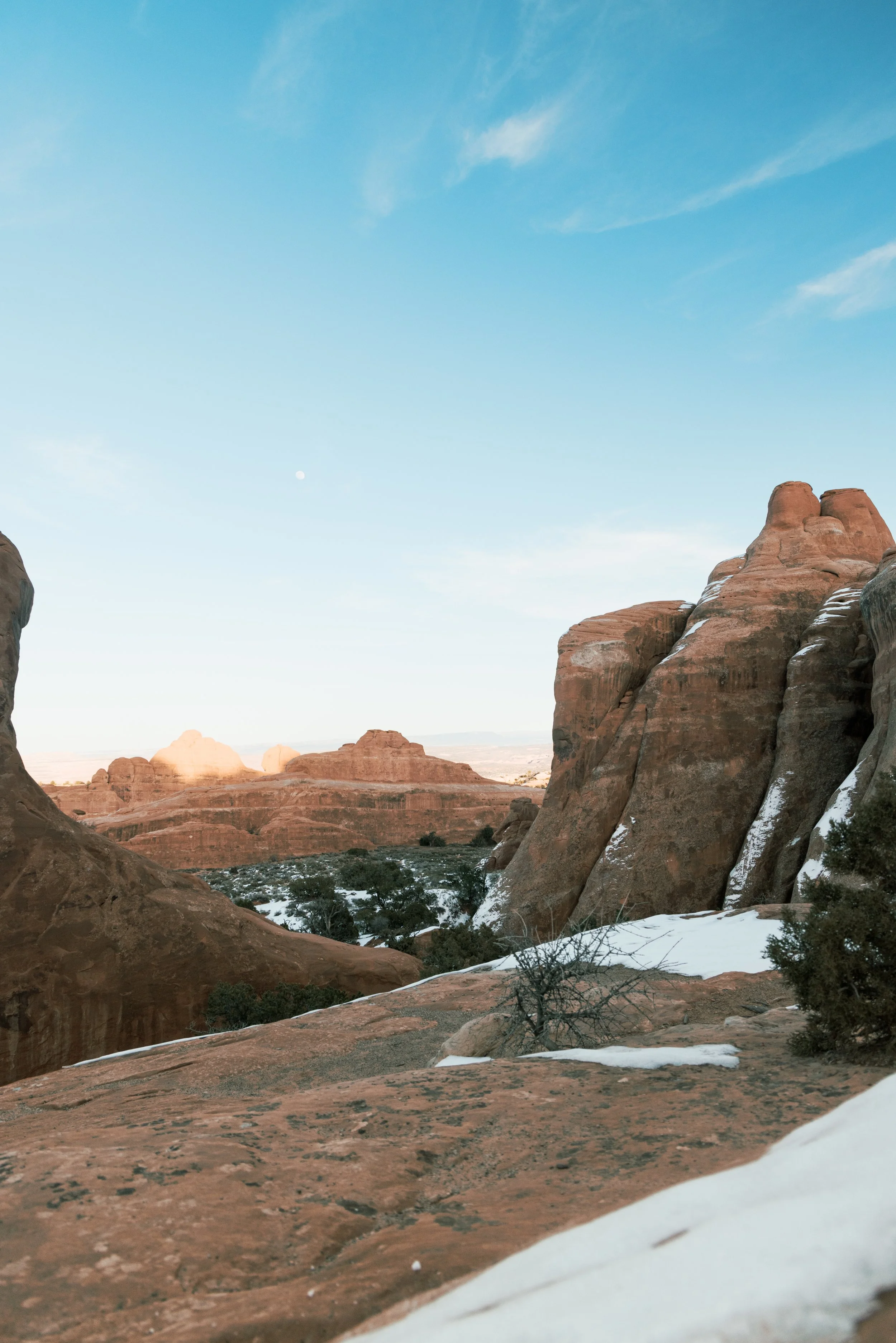 Wide shot of a desert landscape with red rocky formations, sparse vegetation, patches of snow, and a clear blue sky with a few wispy clouds.