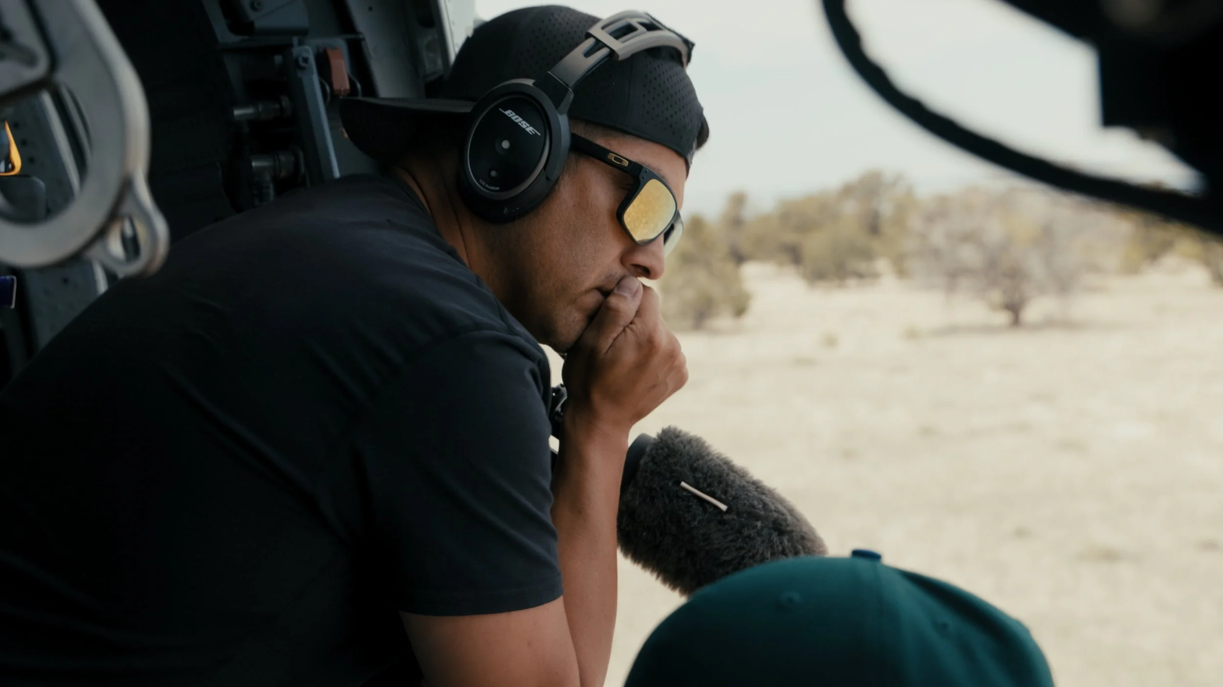 A man wearing a cap, sunglasses, and Bose headphones looking out the window of a helicopter, appearing thoughtful, with a desert landscape in the background.