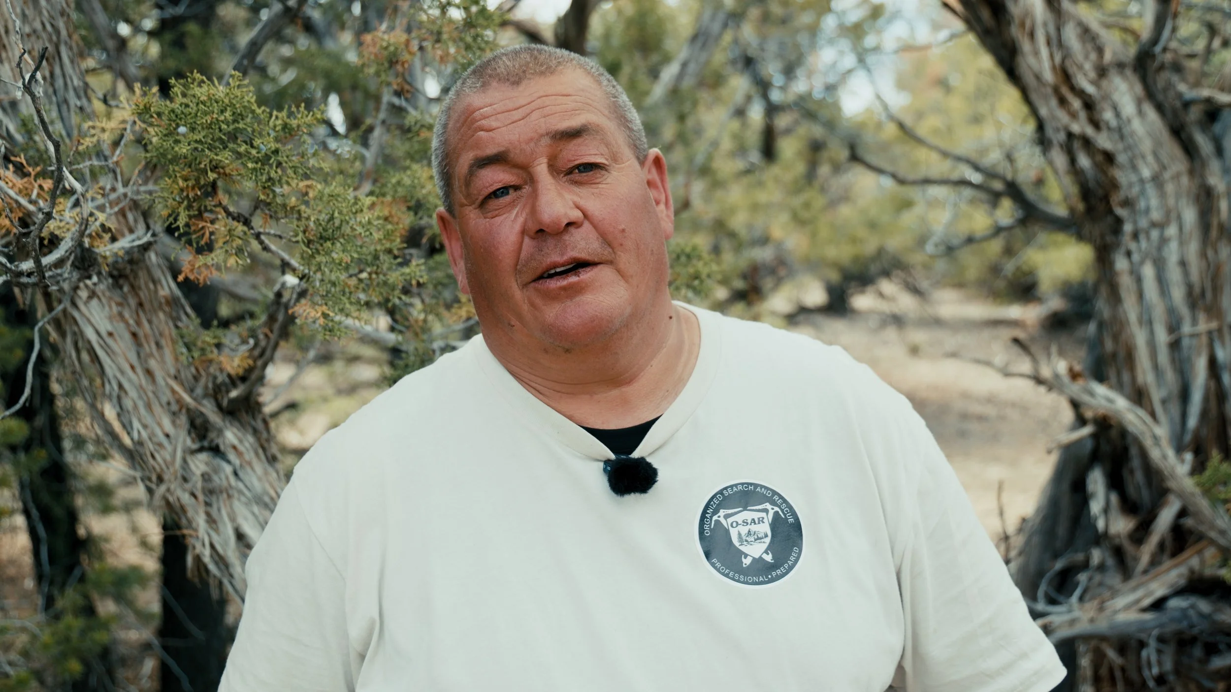 A middle-aged man with short gray hair and a beige T-shirt standing outdoors among trees and shrubs. He is wearing a black clip-on microphone on his shirt and appears to be speaking or explaining something.