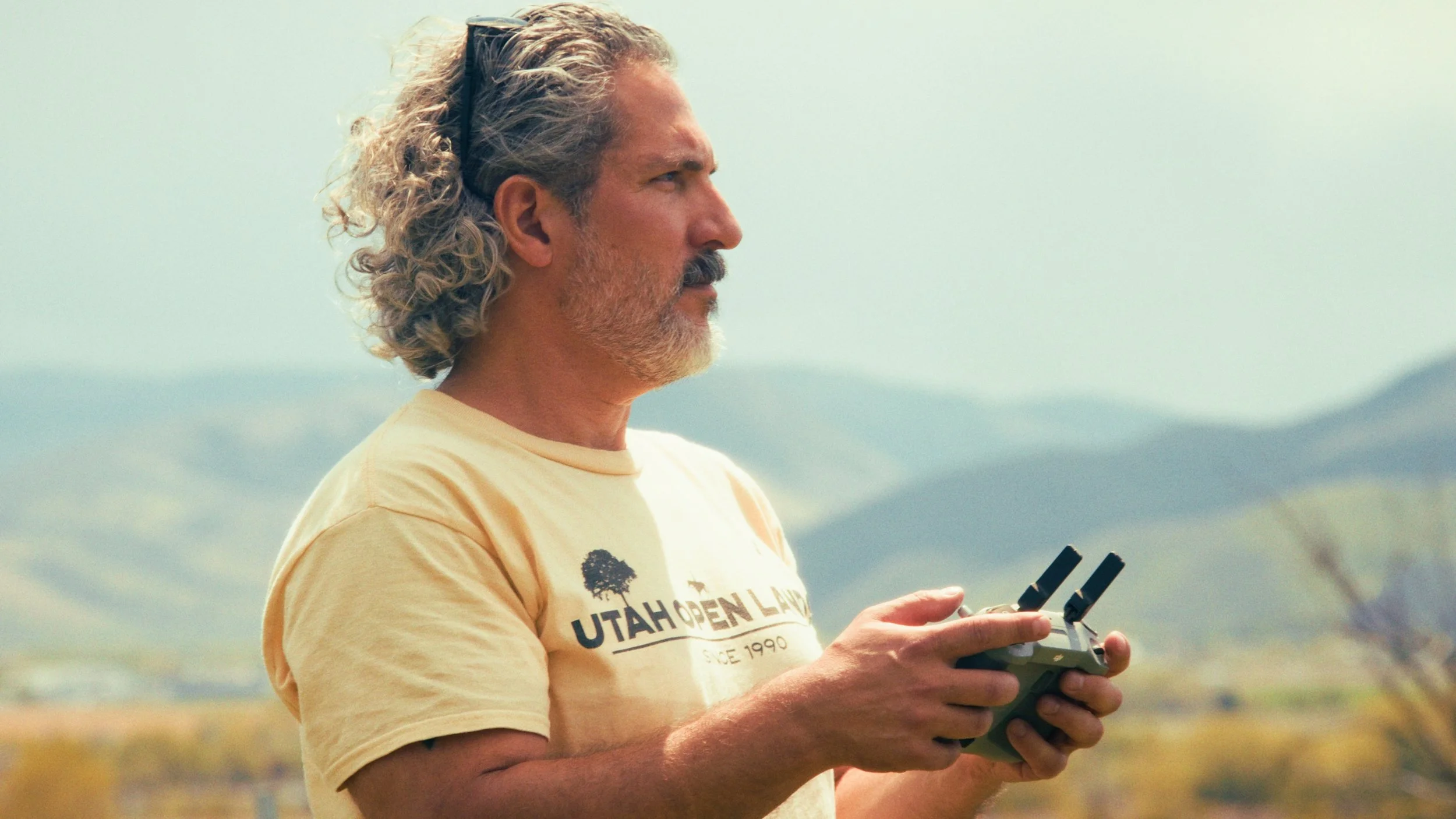 A man with gray curly hair and a beard, wearing a yellow T-shirt with 'Utah Open Land' printed on it, operating a drone controller outdoors with a natural landscape background.