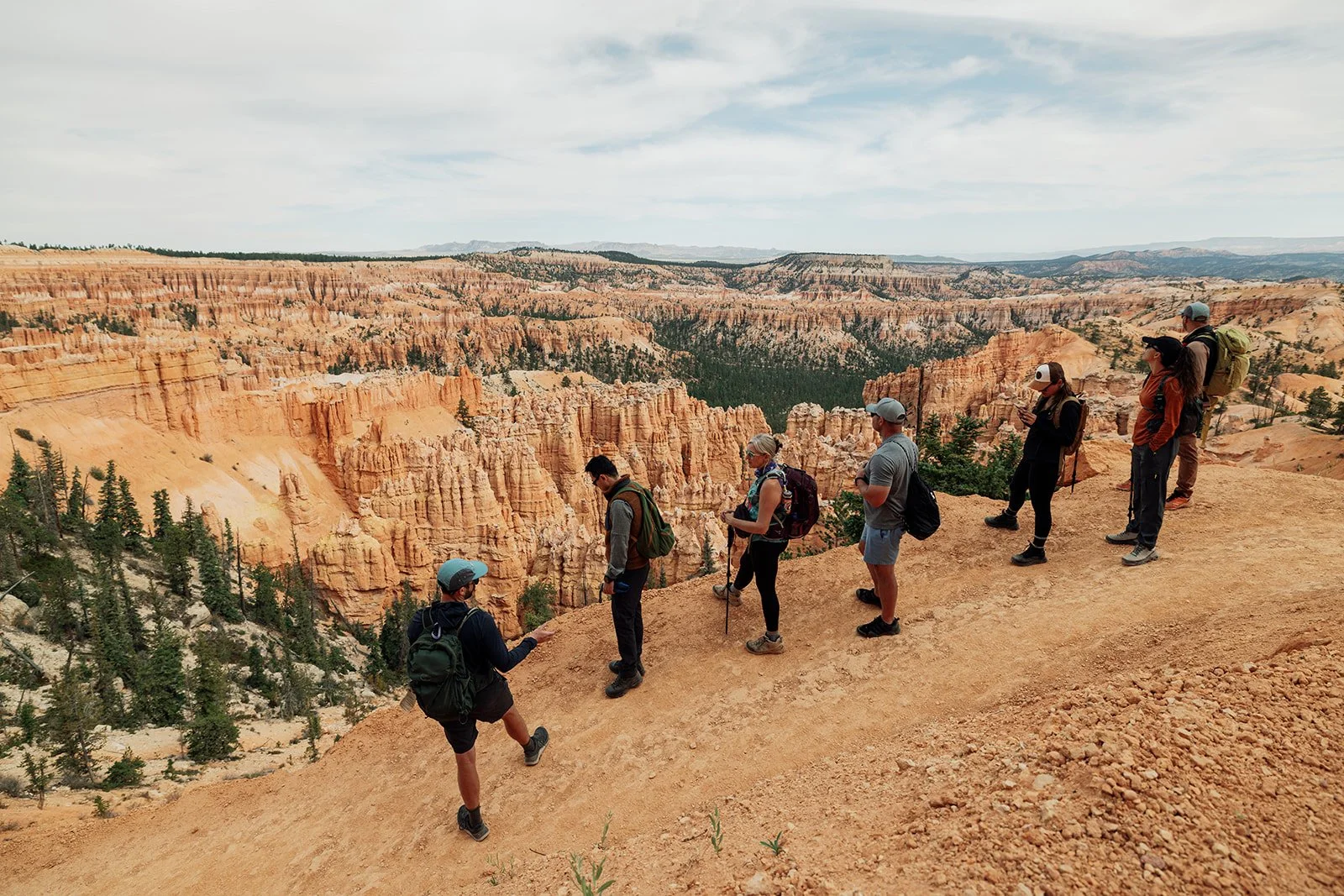 A group of hikers standing on the edge of a canyon overlooking orange rock formations and pine trees, with a cloudy sky overhead.