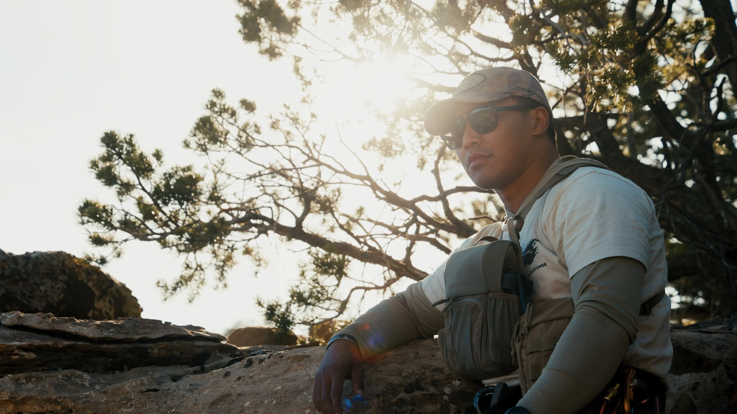 A man in outdoor gear, including sunglasses, a hat, and a backpack, sitting on a rocky terrain with trees and sunlight in the background.