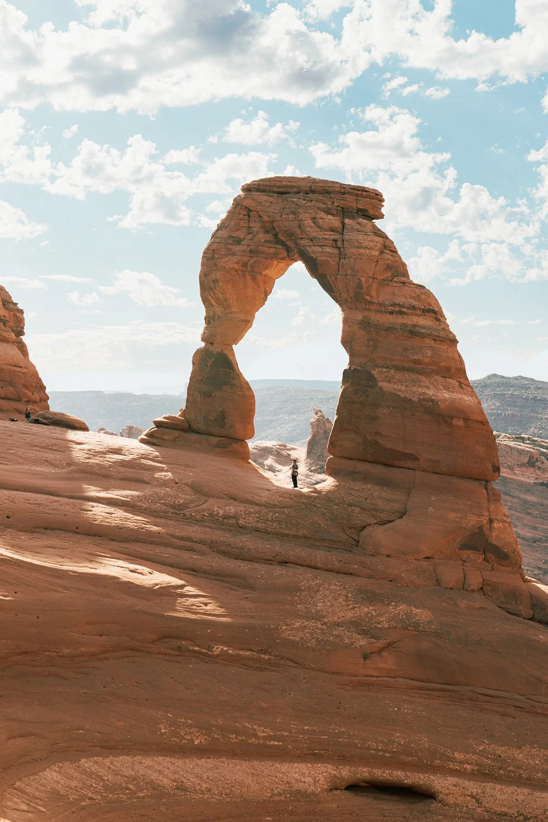 Arches National Park in Utah featuring the Delicate Arch natural sandstone formation with a person standing at its base, under a partly cloudy sky.