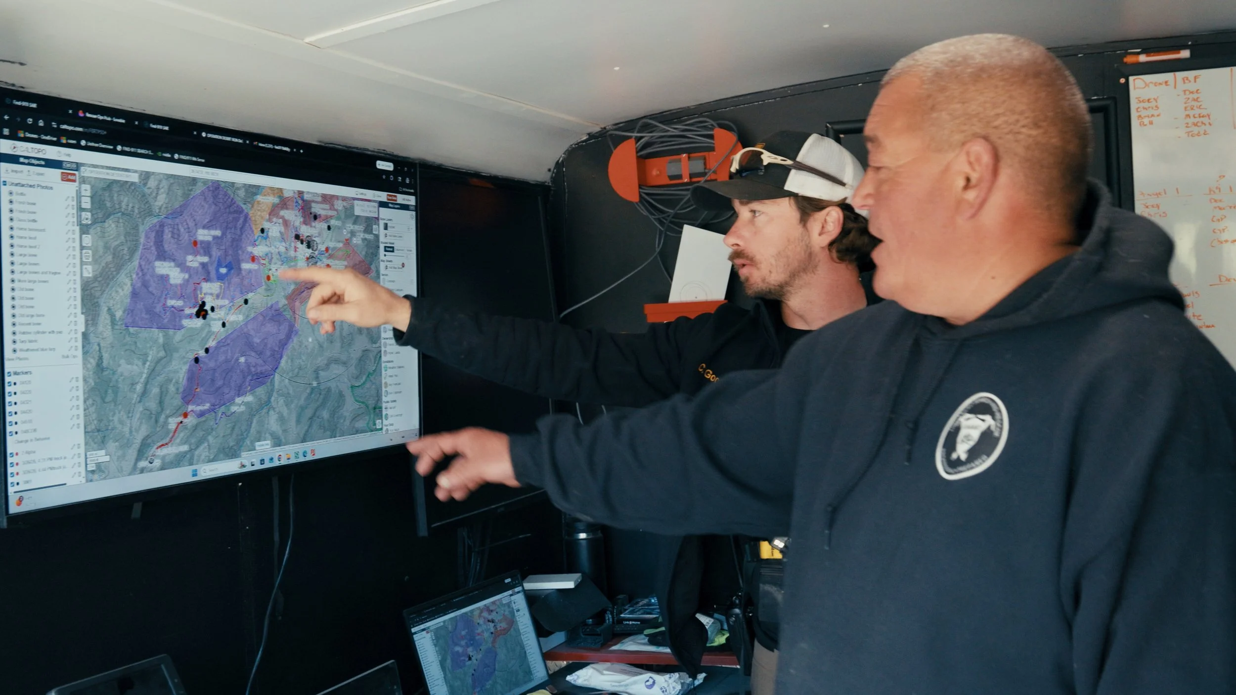 Two men reviewing a map on a large computer screen, pointing and discussing. One is wearing a hat with headlamp, the other is bald and wearing a hoodie. Electronic equipment and notes are visible around them.