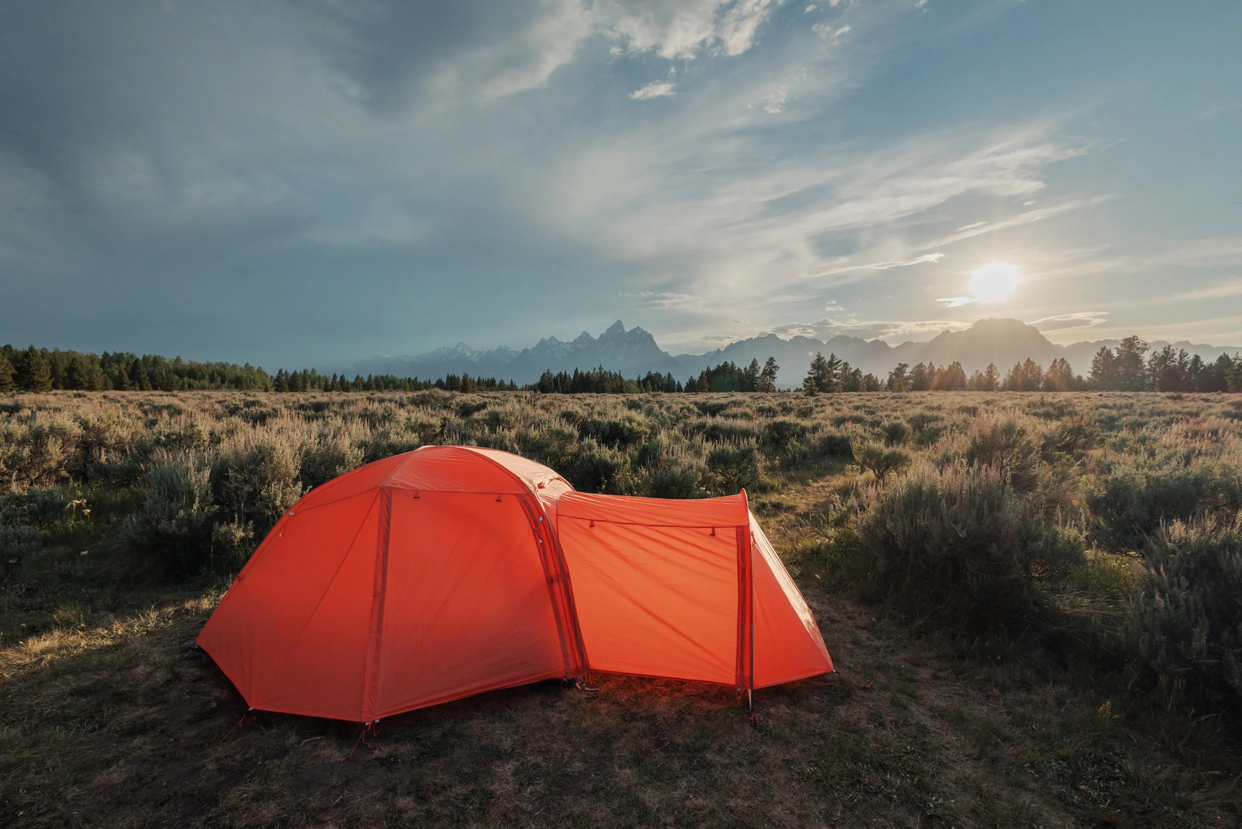 A red tent set up in a grassy field with shrubs, trees, mountains, and the sun setting or rising in the background, under a partly cloudy sky.