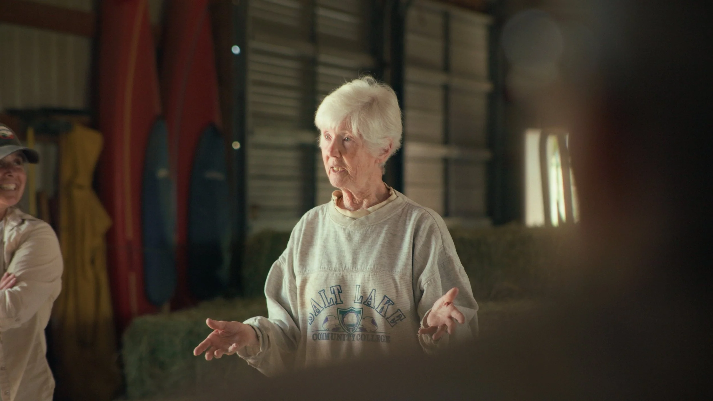 An elderly woman with white hair and a beige sweatshirt standing indoors, talking, with another person partially visible on the left. The background shows colorful kayaks stored on a wall.