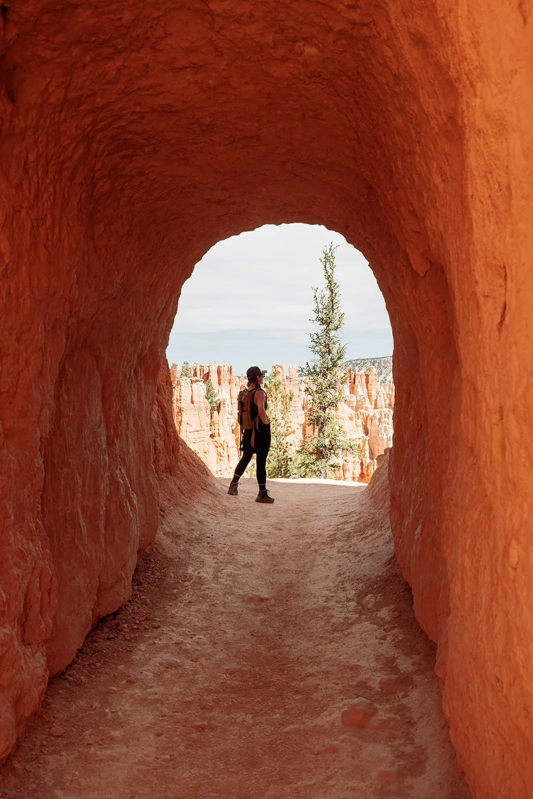 Female hiker with backpack and hat standing in a natural red rock tunnel, looking out at rocky formations and trees in a desert landscape.