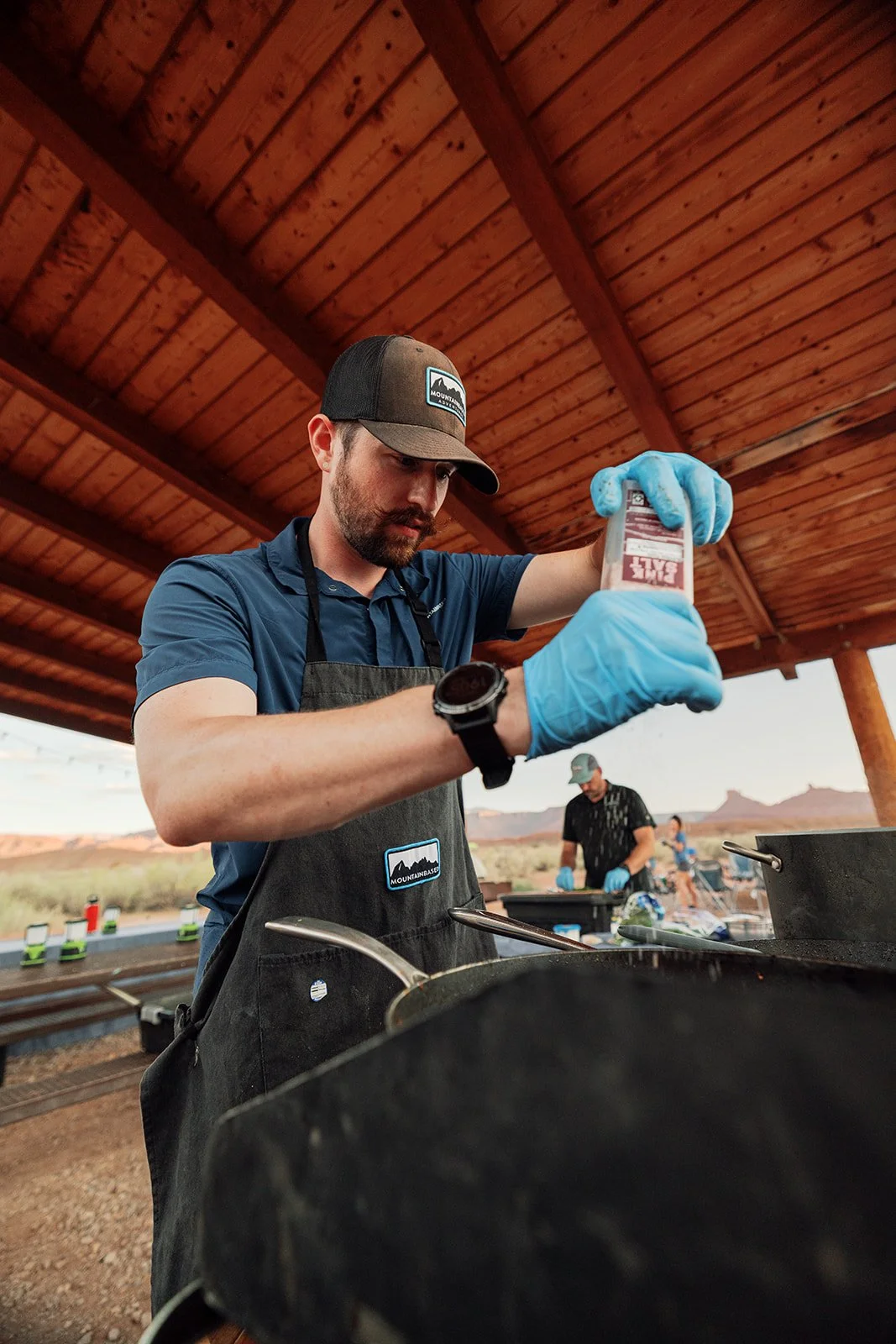 A man in a baseball cap and apron wearing blue gloves is pouring a bottle of sauce into a black grill. Other people are visible in the background under a wooden pavilion with a desert landscape in the distance.