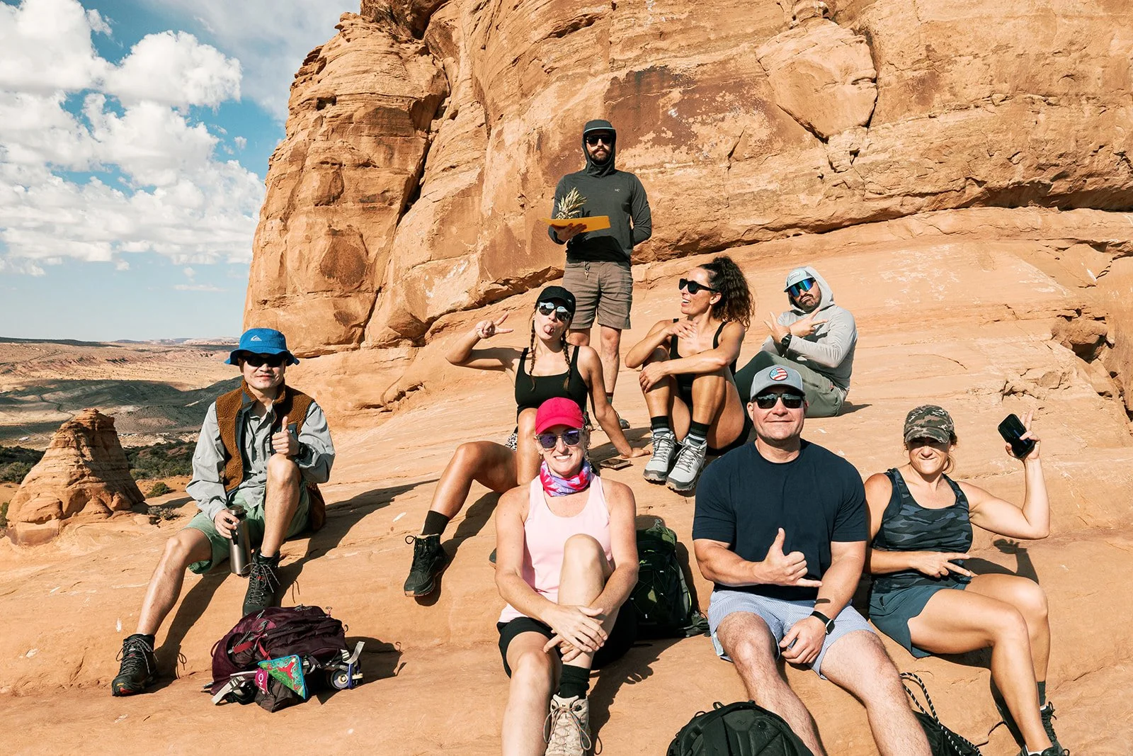 A group of nine people sitting and standing on a large rock formation in a desert landscape with orange cliffs, blue sky, and scattered clouds.