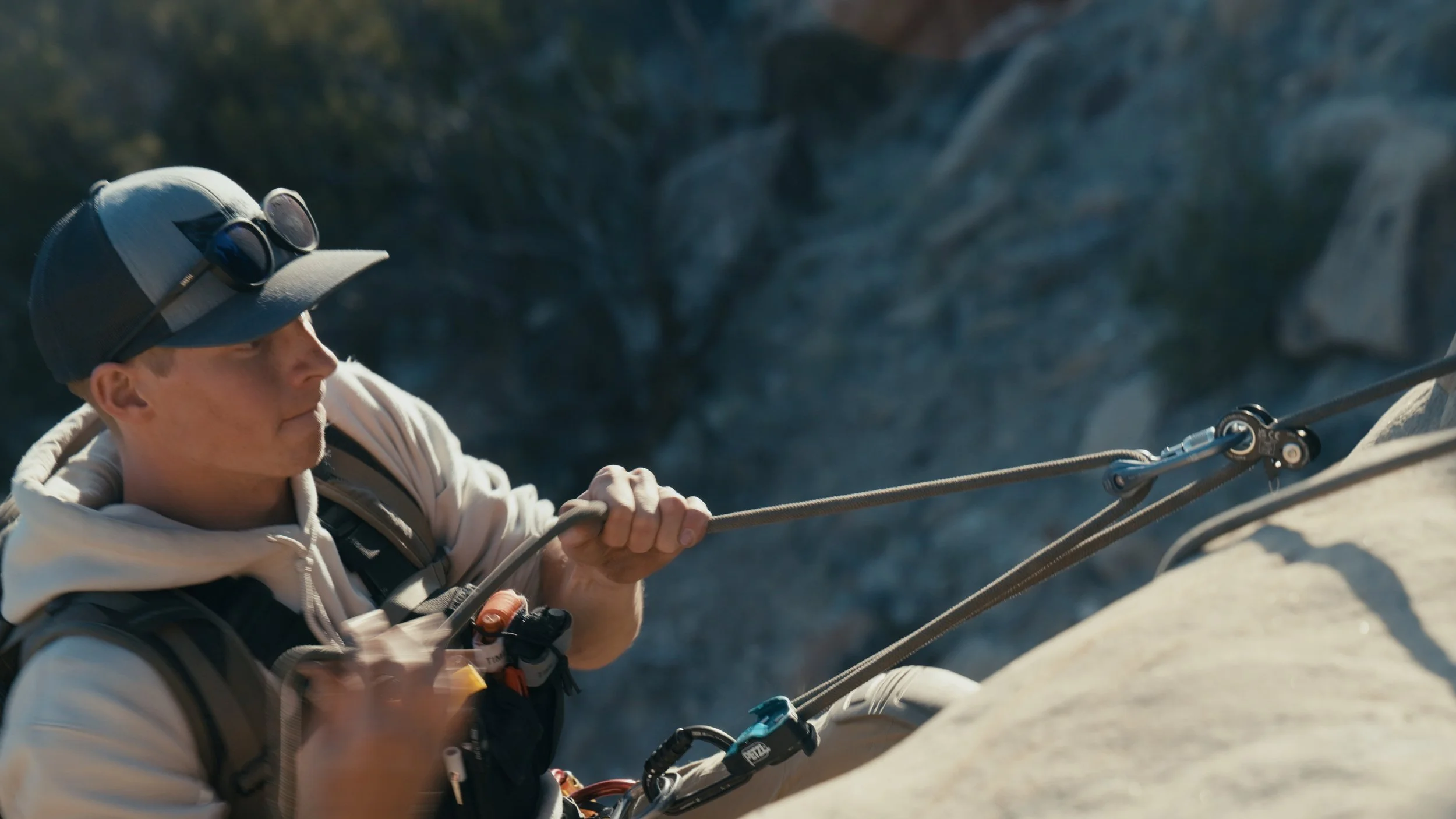 A man wearing a baseball cap with goggles on top, white hoodie, and climbing harness, is rock climbing on a steep outdoor rock face, using a safety rope and equipment.