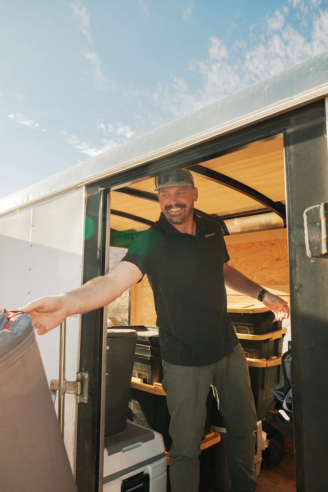 Man smiling and reaching outside a mobile food or camping cart, dressed in black shirt and cap, with storage bins inside and outdoors under a cloudy sky.