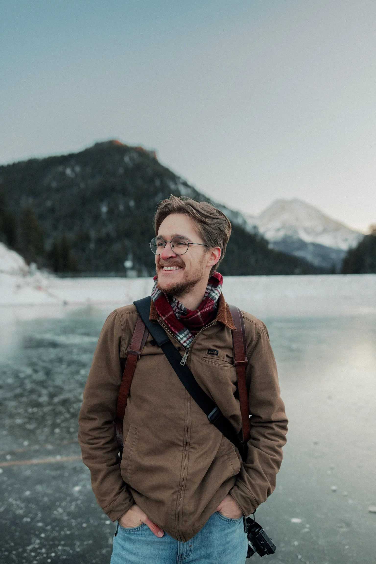 A young man with glasses and a beard standing outdoors near a body of water, wearing a brown jacket and a plaid scarf, with mountains in the background.
