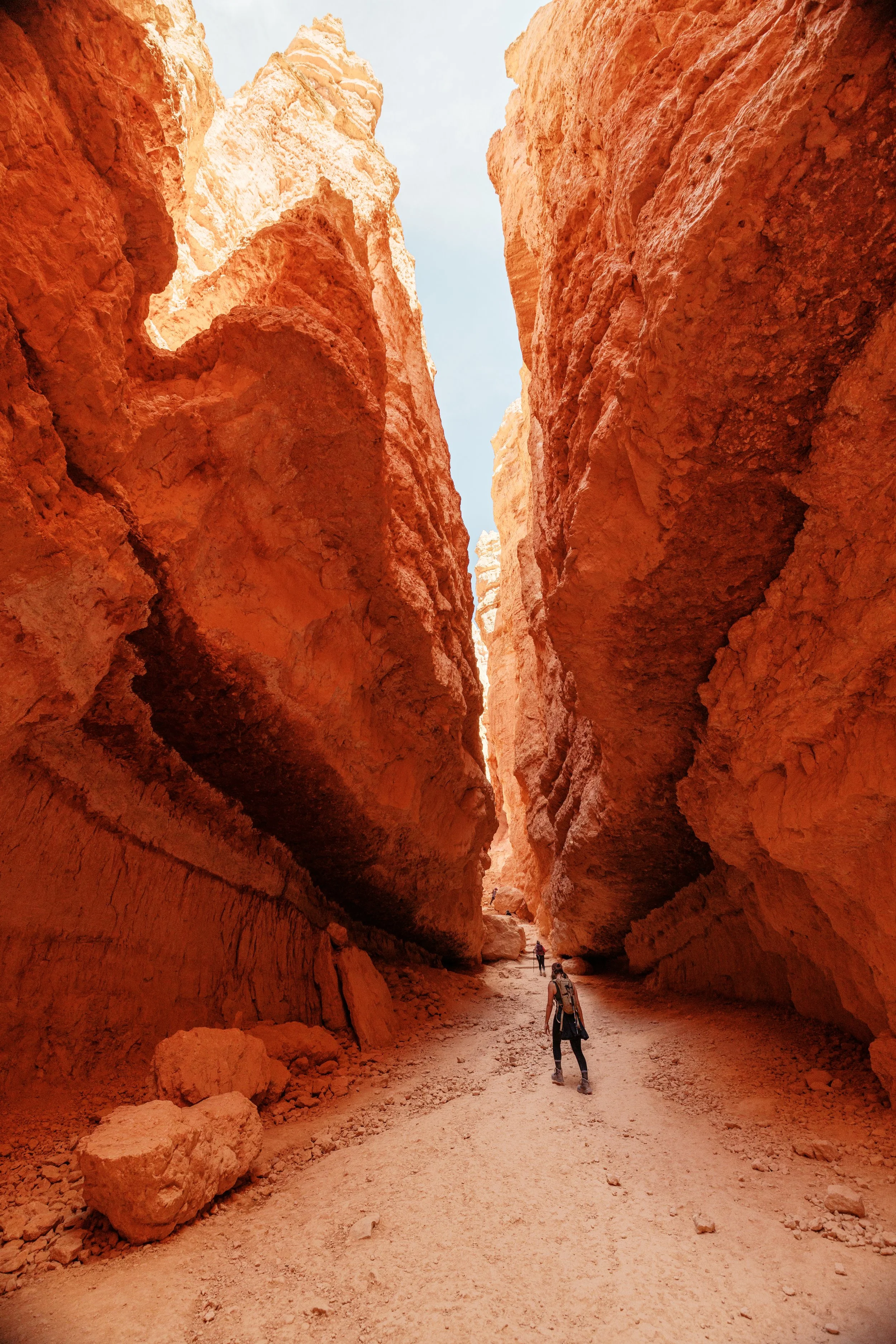People hiking through a narrow canyon with tall, red rock walls on either side, under a light blue sky.