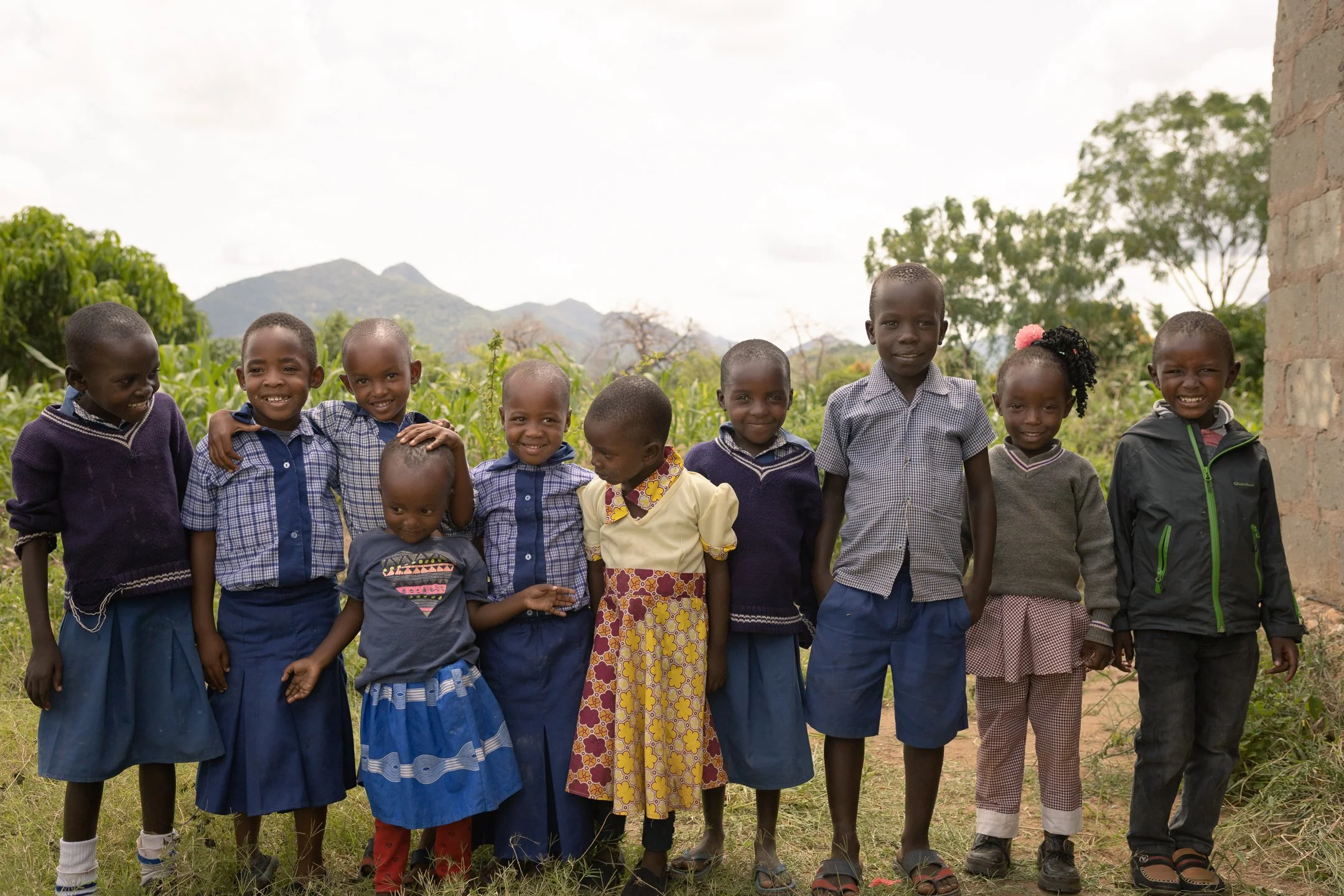 Group of ten children standing together outdoors with greenery and mountains in the background, smiling at the camera, wearing school uniforms and casual clothes.