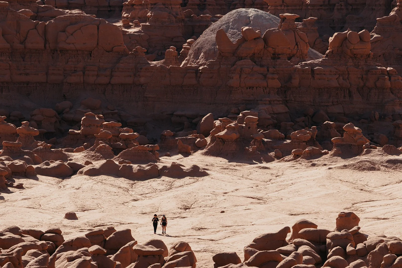 Two hikers walking across a desert landscape with rock formations and cliffs in the background.