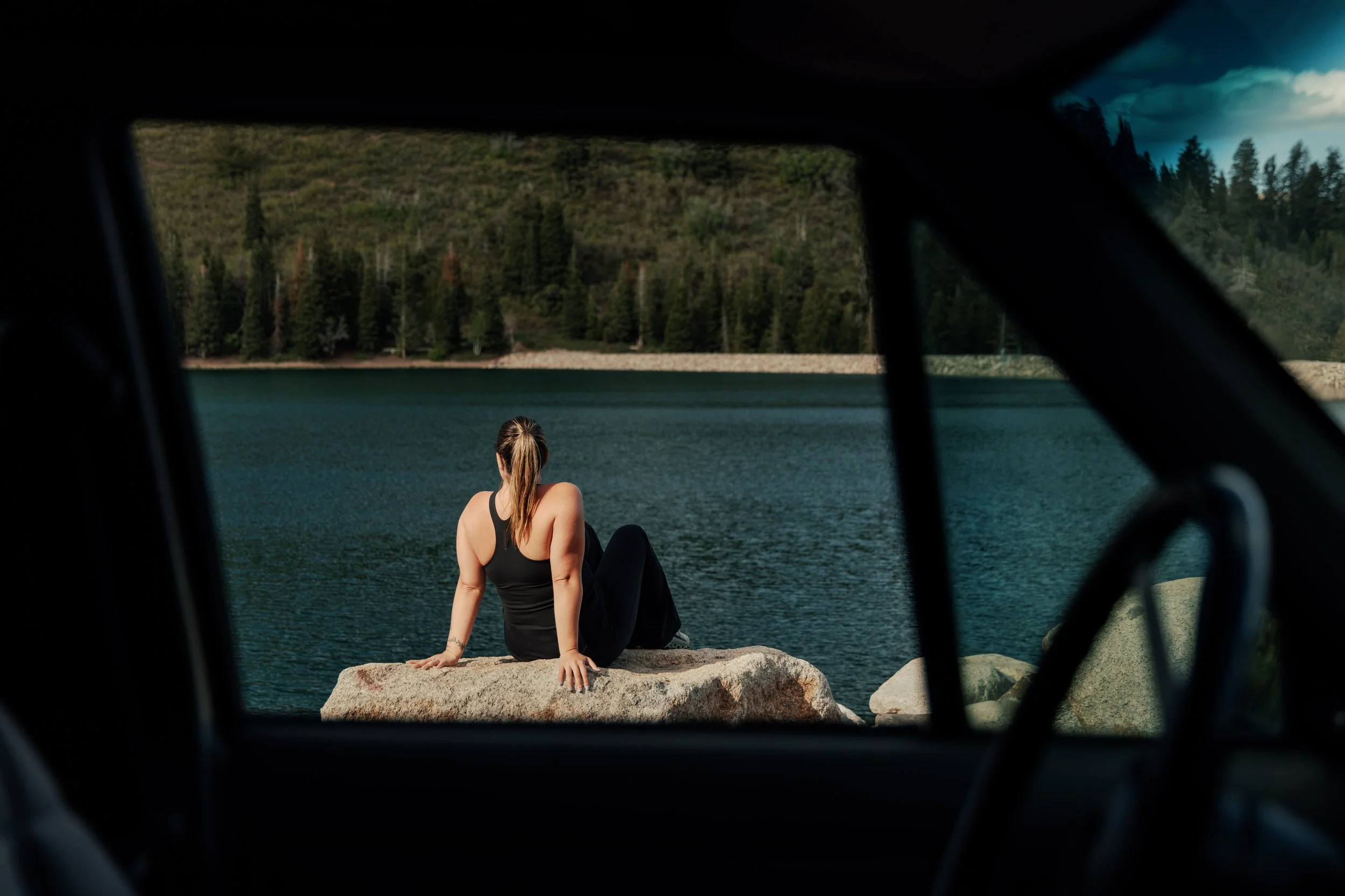 A woman sitting on a rock near a body of water, viewed through the window of a vehicle with trees and a cloudy sky in the background.