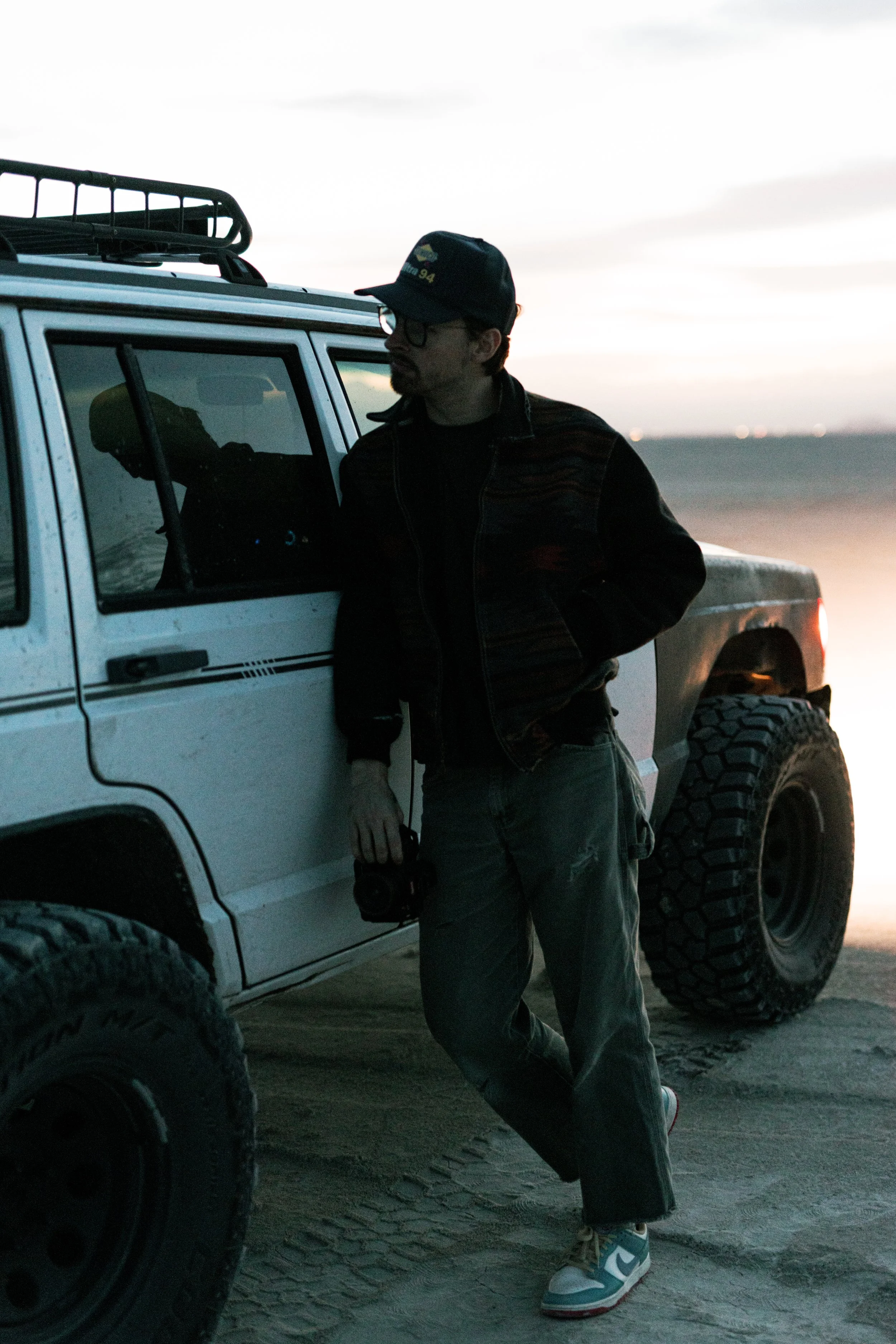 Person wearing glasses, a black cap, black jacket, baggy green pants, and sneakers standing next to a white off-road vehicle during sunset or dawn.