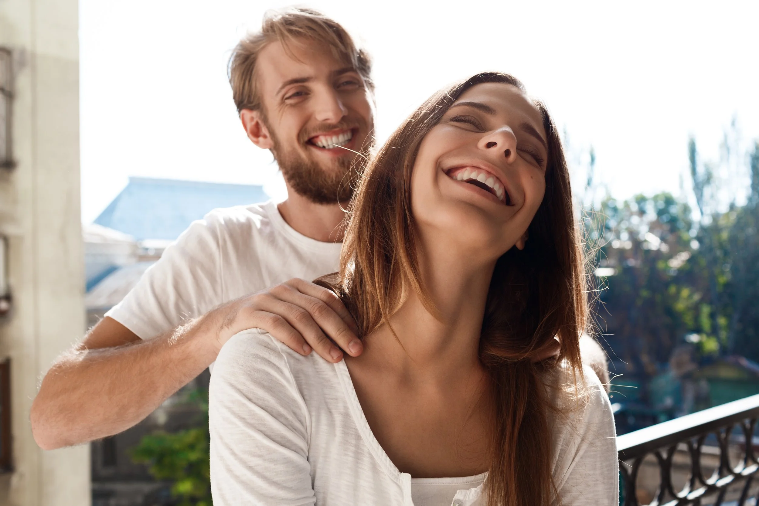A man giving a piggyback ride to a woman on a balcony, both smiling and enjoying the moment with a sunny outdoor background.