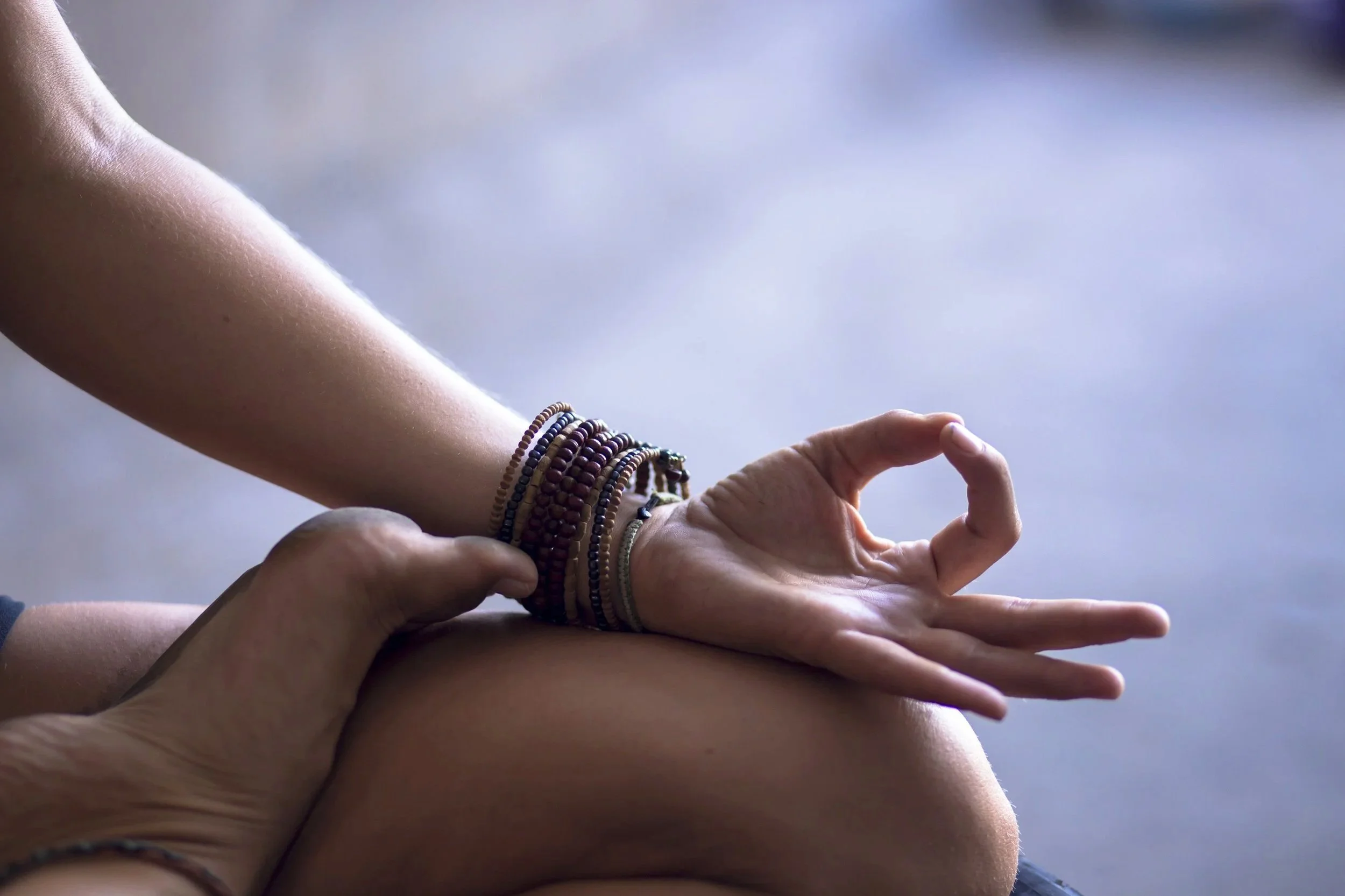 Close-up of a person’s hand resting on their knee, with the thumb and index finger touching, in a gesture of meditation or yoga. The person is wearing several beaded bracelets on their wrist.