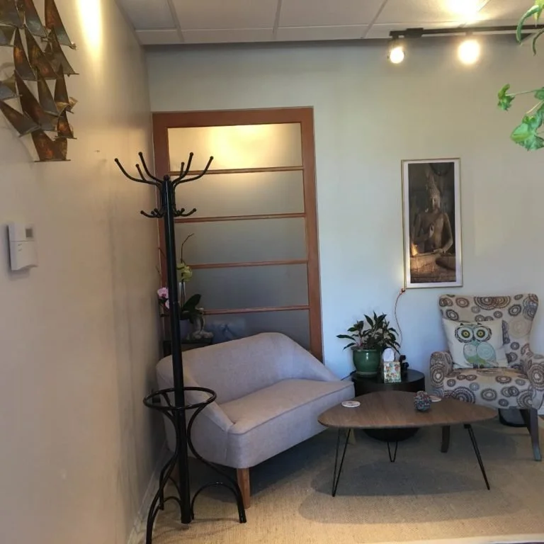 Living room with a beige couch, a patterned armchair, a small wooden coffee table, and a green potted plant. There is a wall art of a seated Buddha and a wooden room divider in the background.
