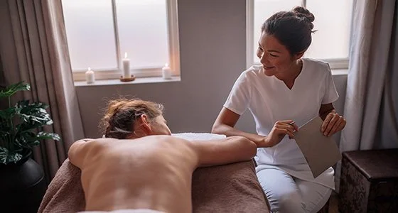 A medical professional in white scrubs consulting with a patient lying face down on a treatment table in a brightly lit room.