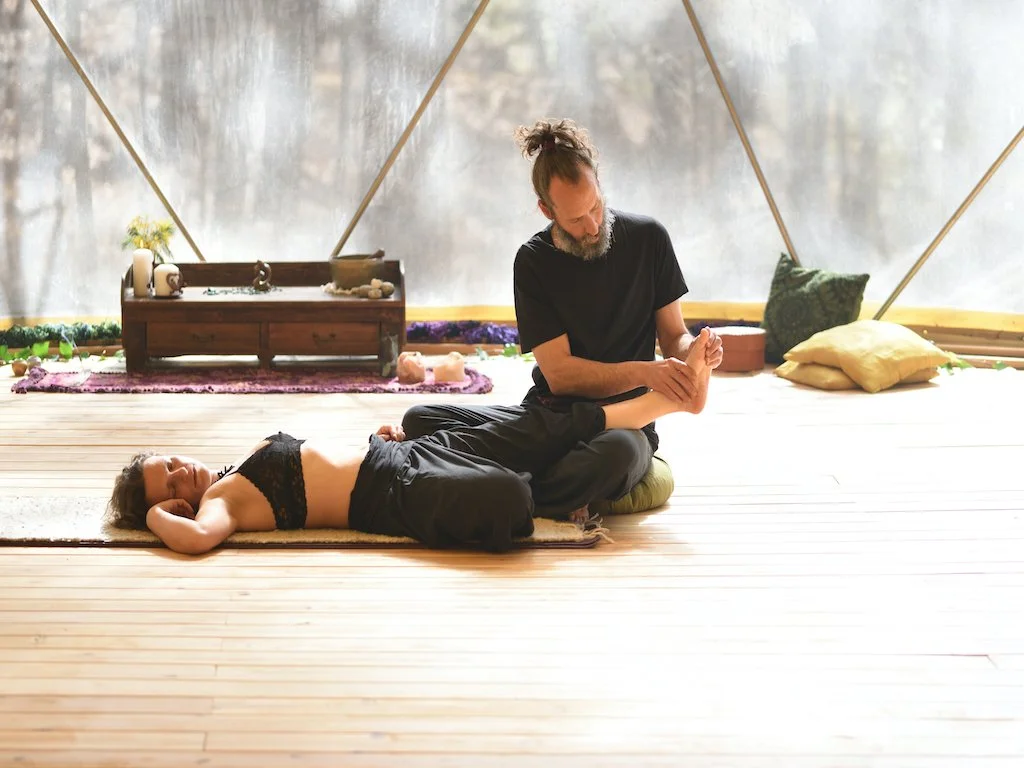 A woman lying on her back on a yoga mat with her eyes closed, receiving a foot massage from a man sitting beside her, in a cozy, natural setting with wooden floors and soft cushions.