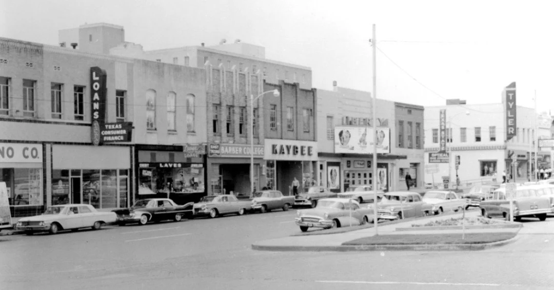 Black and white photo of a small downtown street with cars parked along the curb and various storefronts, including a loan office, a beauty college, and a tailor shop.