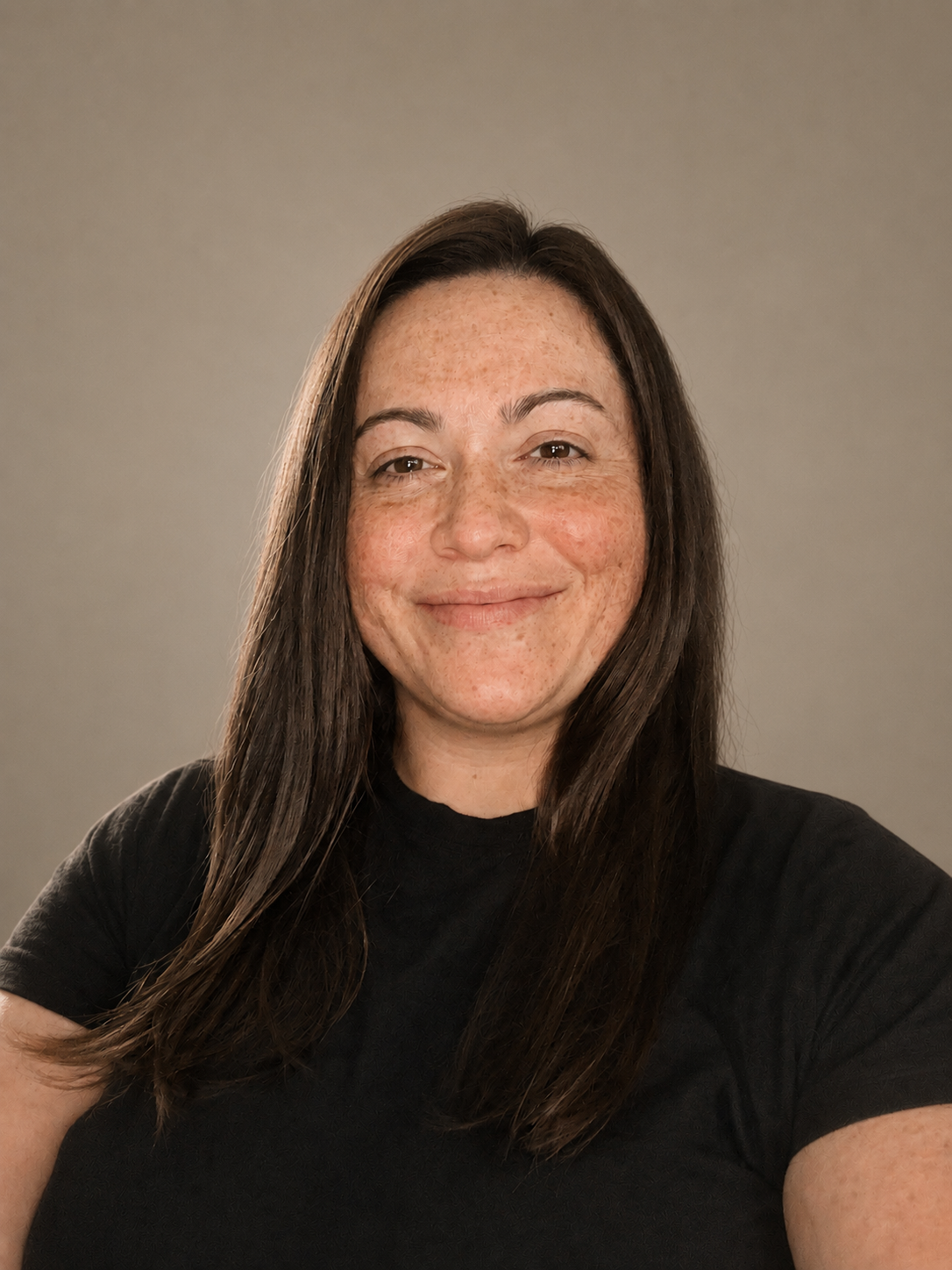 A woman with brown hair and freckles is smiling at the camera, wearing a black shirt against a plain background.