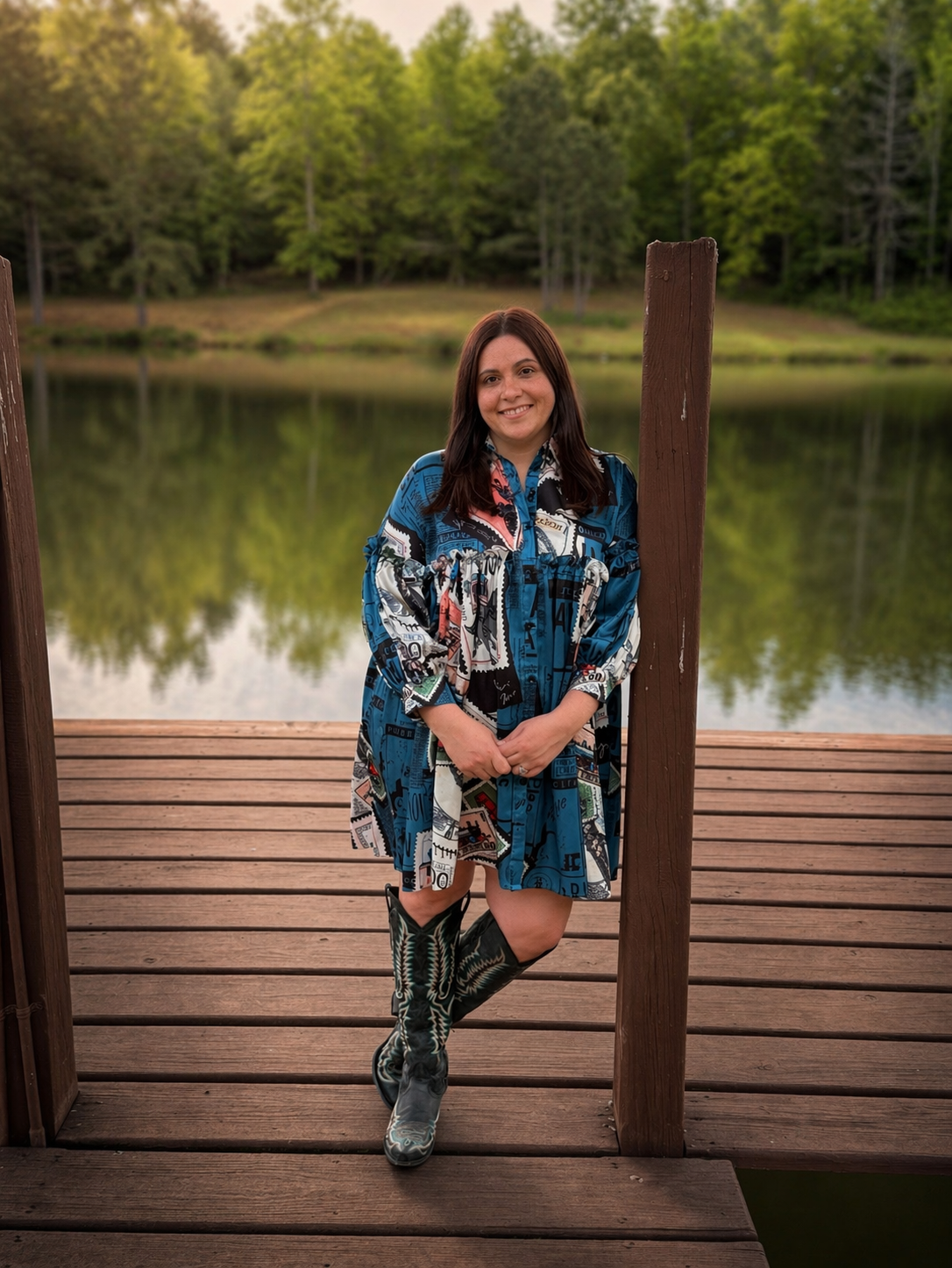 A woman standing on a wooden dock by a lake, with green trees in the background, wearing a colorful patterned dress and cowboy boots, smiling at the camera.