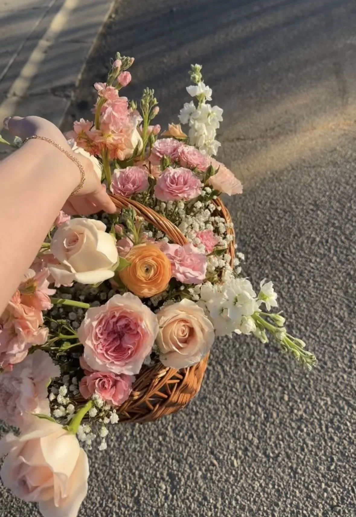 A hand holding a basket of assorted pink, white, and peach flowers on a paved sidewalk during sunlight.