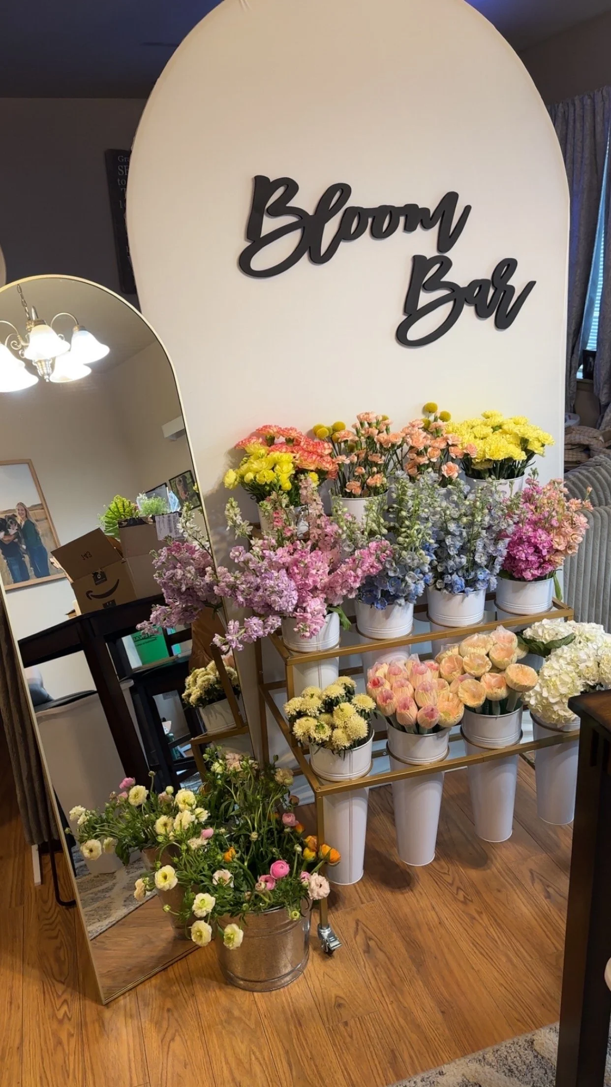 Display of colorful flowers in white pots on a wooden rack and in a large metal bucket, with a decorative mirror and floral-themed sign reading 'Bloom Bar' in the background.