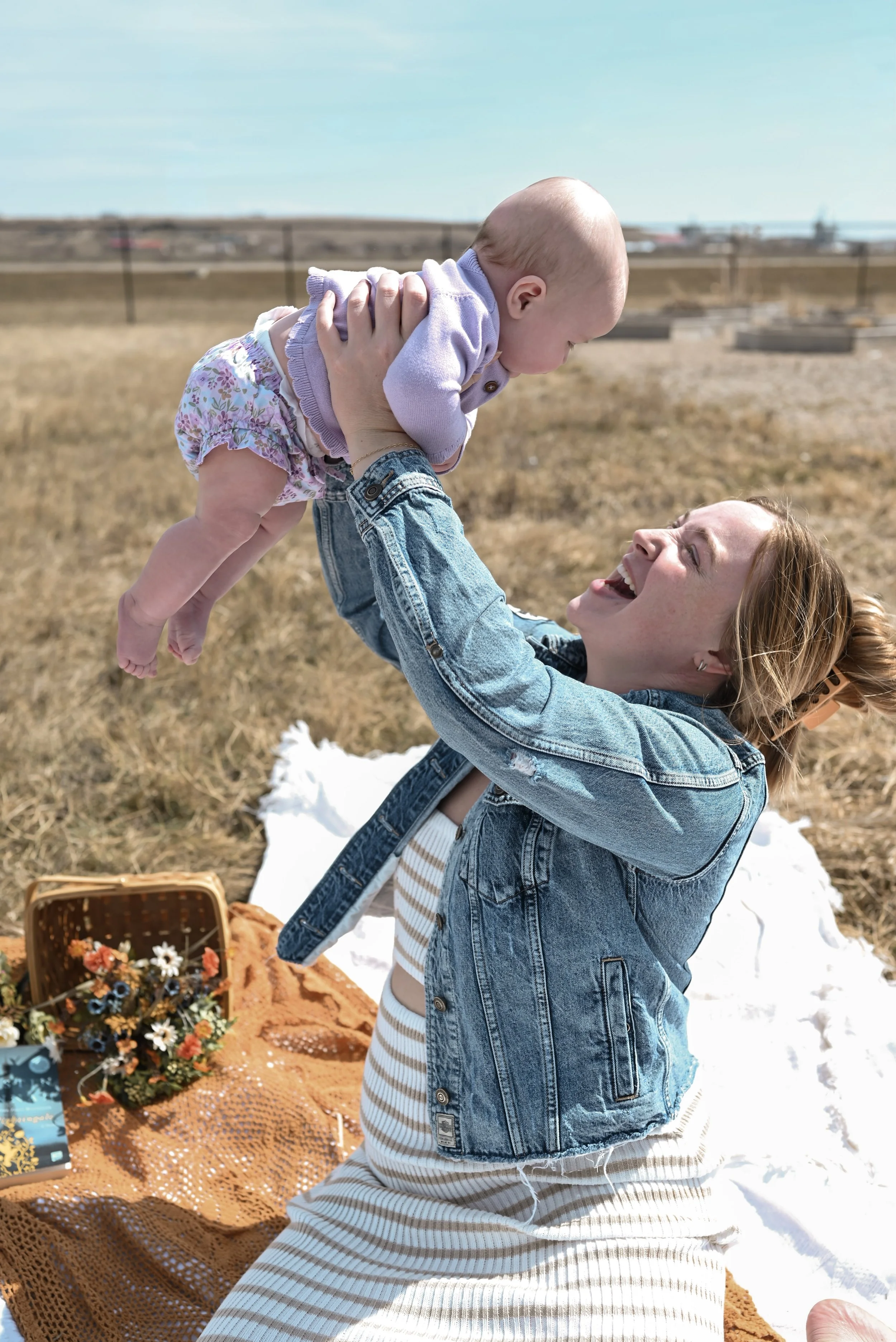 A woman and a baby joyfully playing together outdoors on a sunny day, with a basket of flowers and a blanket nearby.