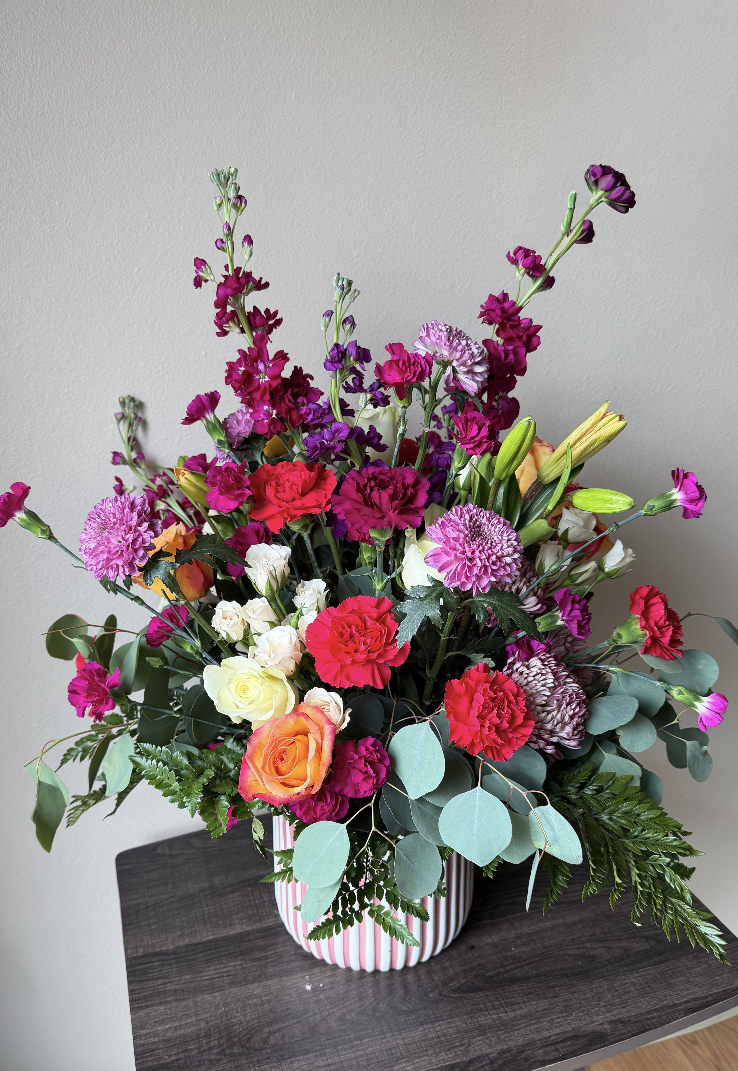 A large floral arrangement in a white and pink striped vase with pink, white, yellow, orange, and purple flowers, and green foliage, placed on a dark wooden table against a plain white wall.