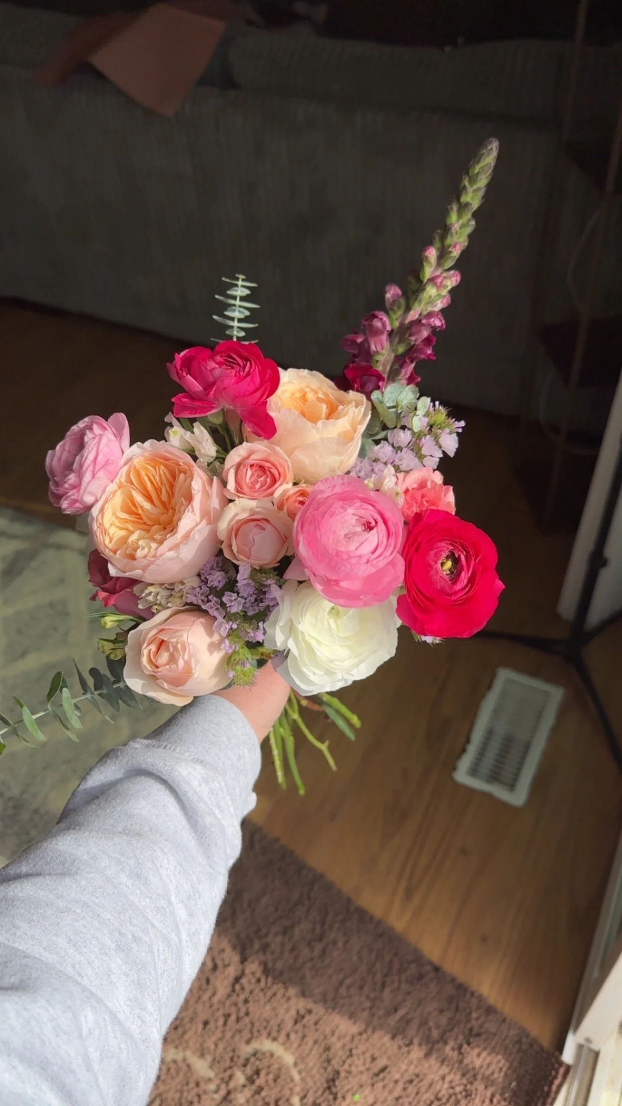 A person holding a bouquet of mixed pink, peach, and white flowers with some purple accent flowers and greenery, in a room with a gray couch and patterned carpet.