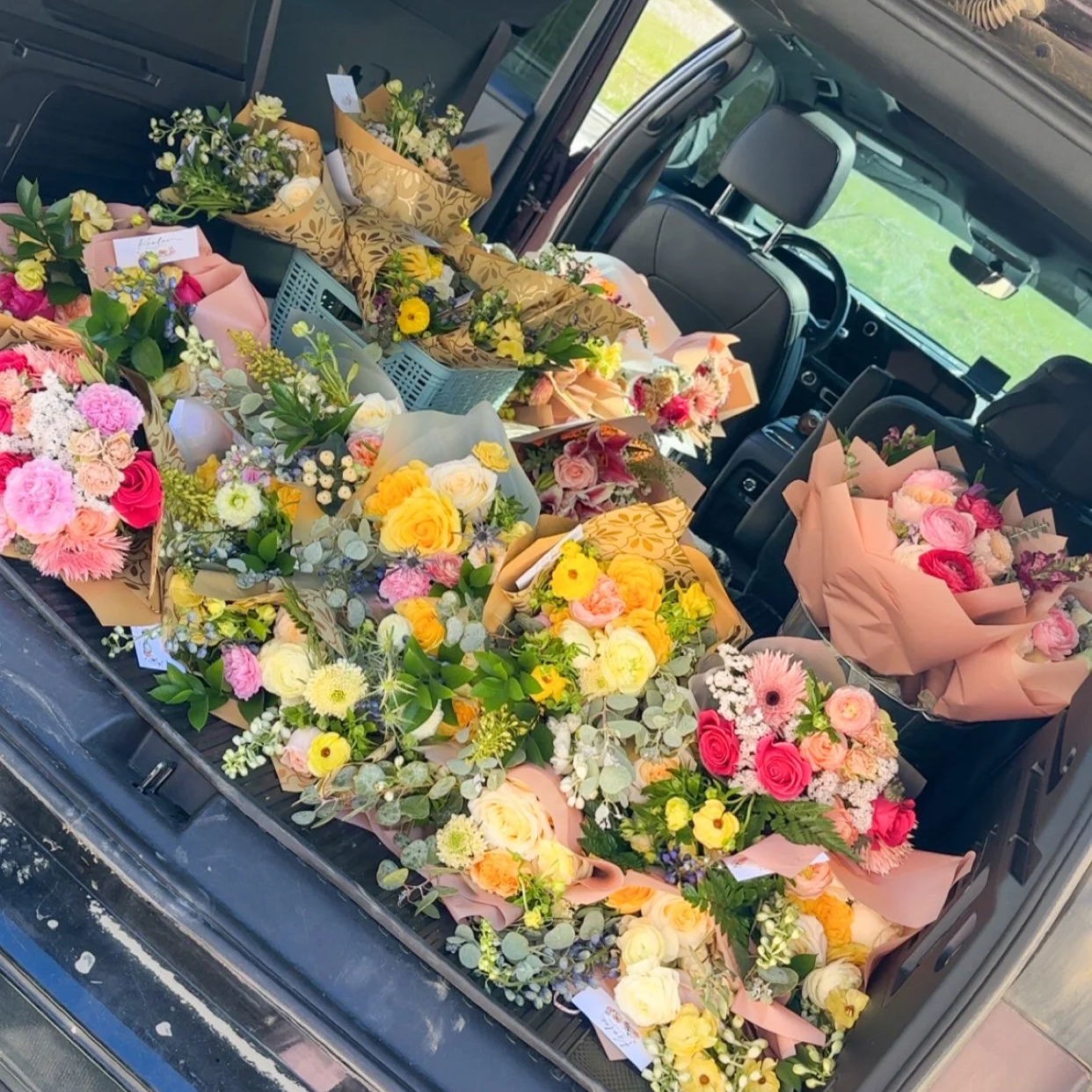 The back of a car filled with numerous colorful flower bouquets, including roses, peonies, and other mixed flowers, wrapped in various floral wrapping paper.