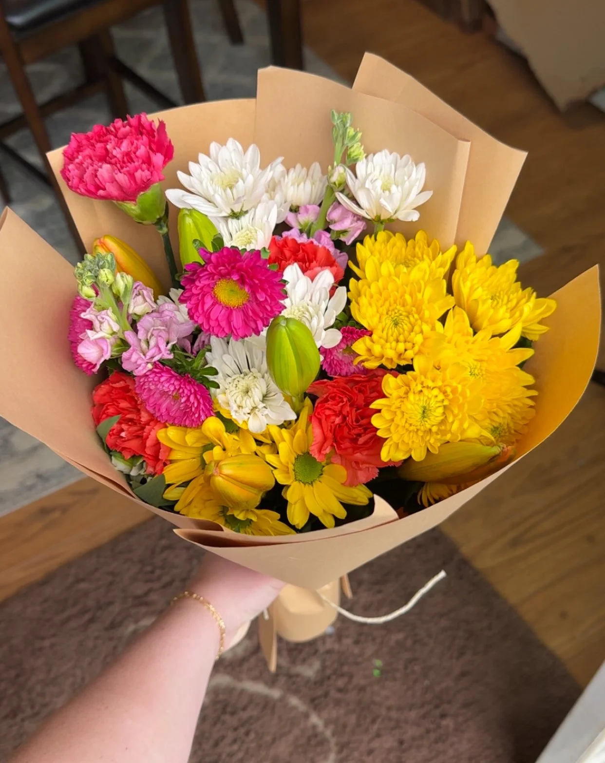 A colorful bouquet of flowers wrapped in light brown paper, featuring pink, white, yellow, and red blossoms, held by a person with a bracelet on their wrist.