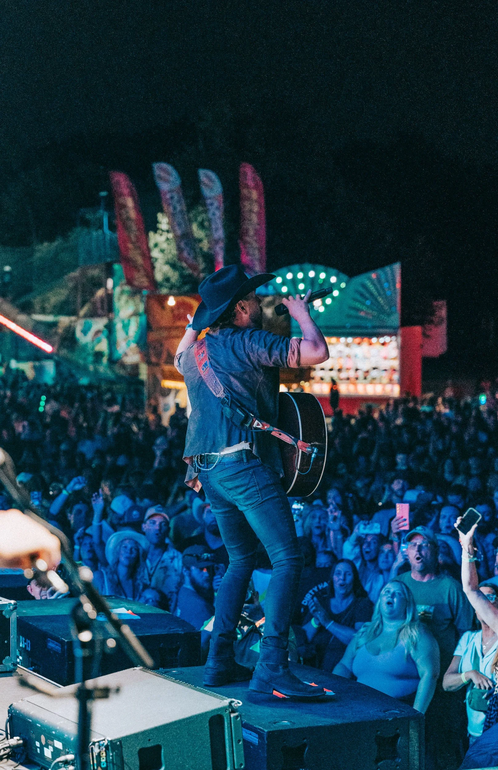 A country singer performing on stage at night, wearing a cowboy hat and playing guitar, in front of a large crowd at a fair or carnival with brightly lit amusement rides.