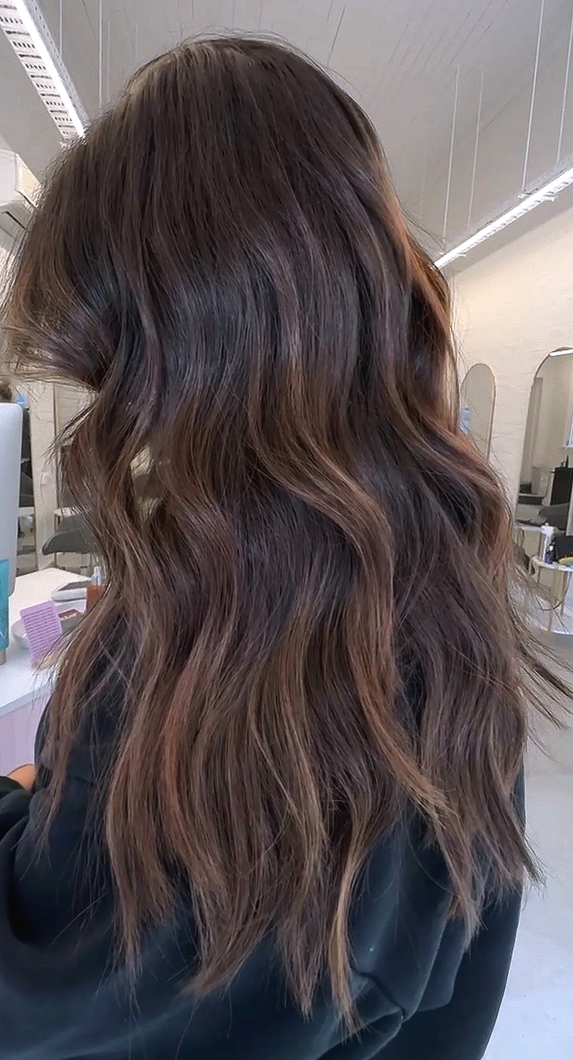 A person with long, wavy brown hair with subtle highlights in a salon or similar setting.